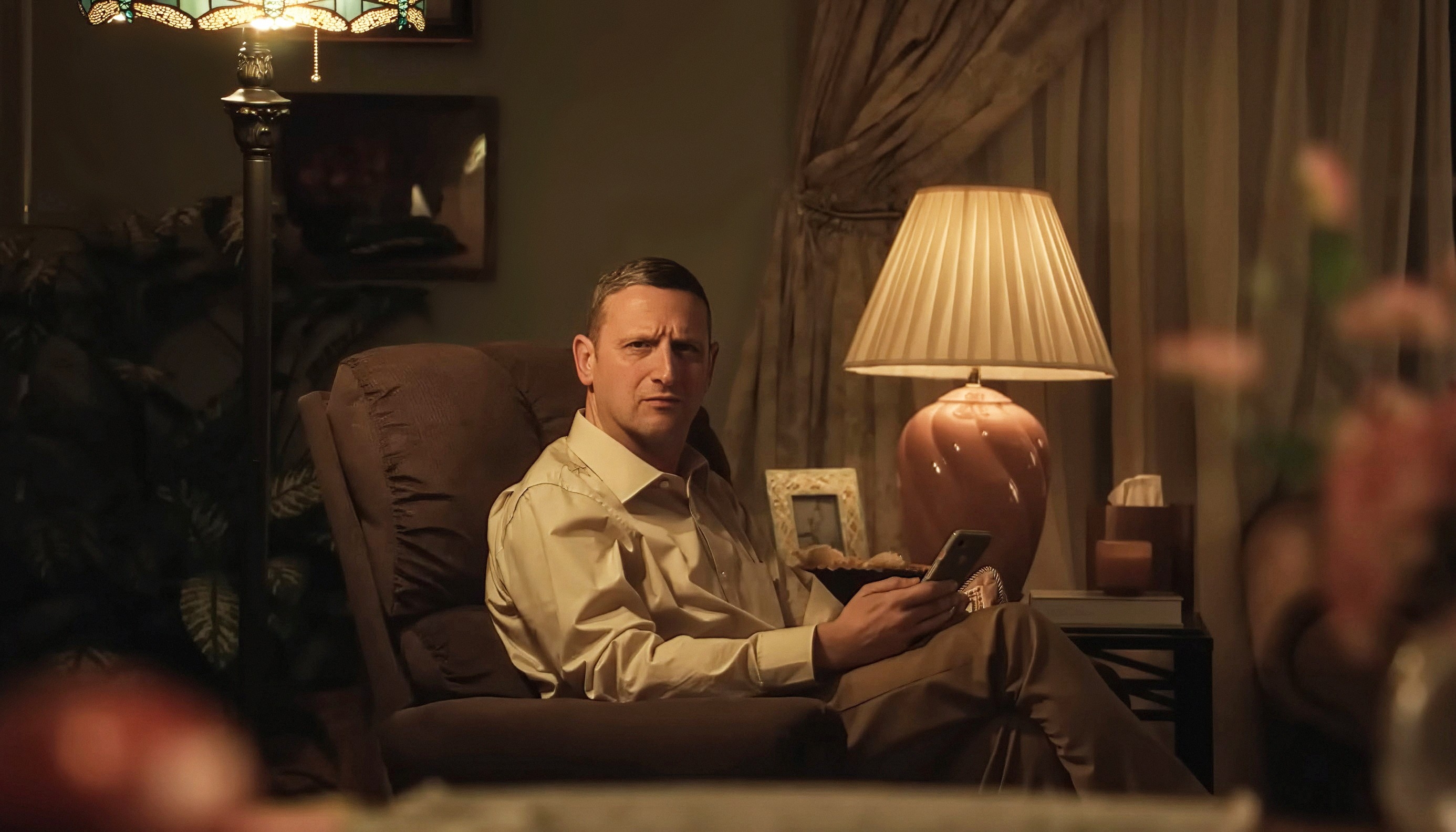 A person sits in an armchair, looking confused while holding a book. The room is warmly lit with a table lamp and framed photos in the background