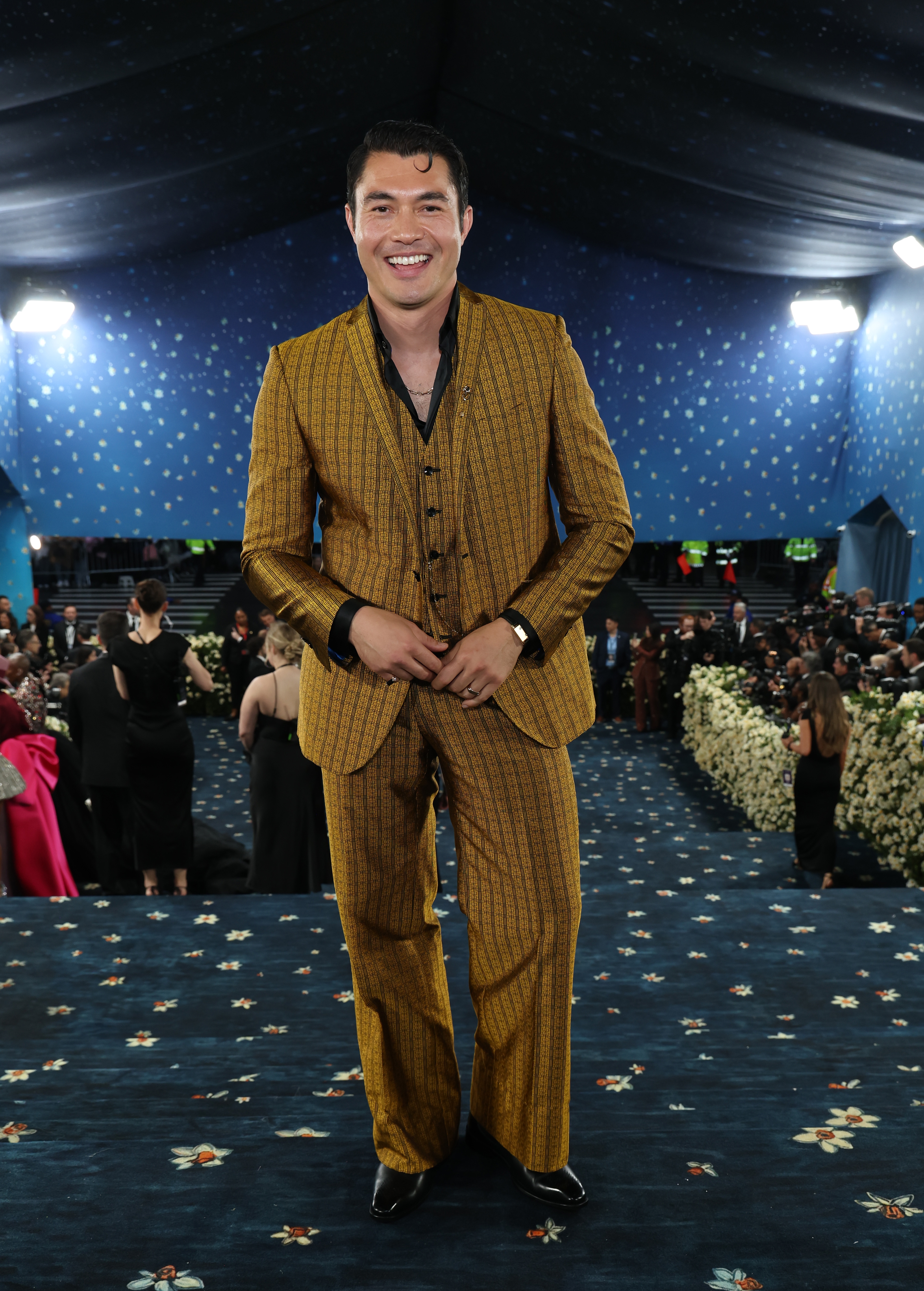Man on a decorative carpet wearing a tailored striped suit with a waistcoat, smiling and posing at a formal event