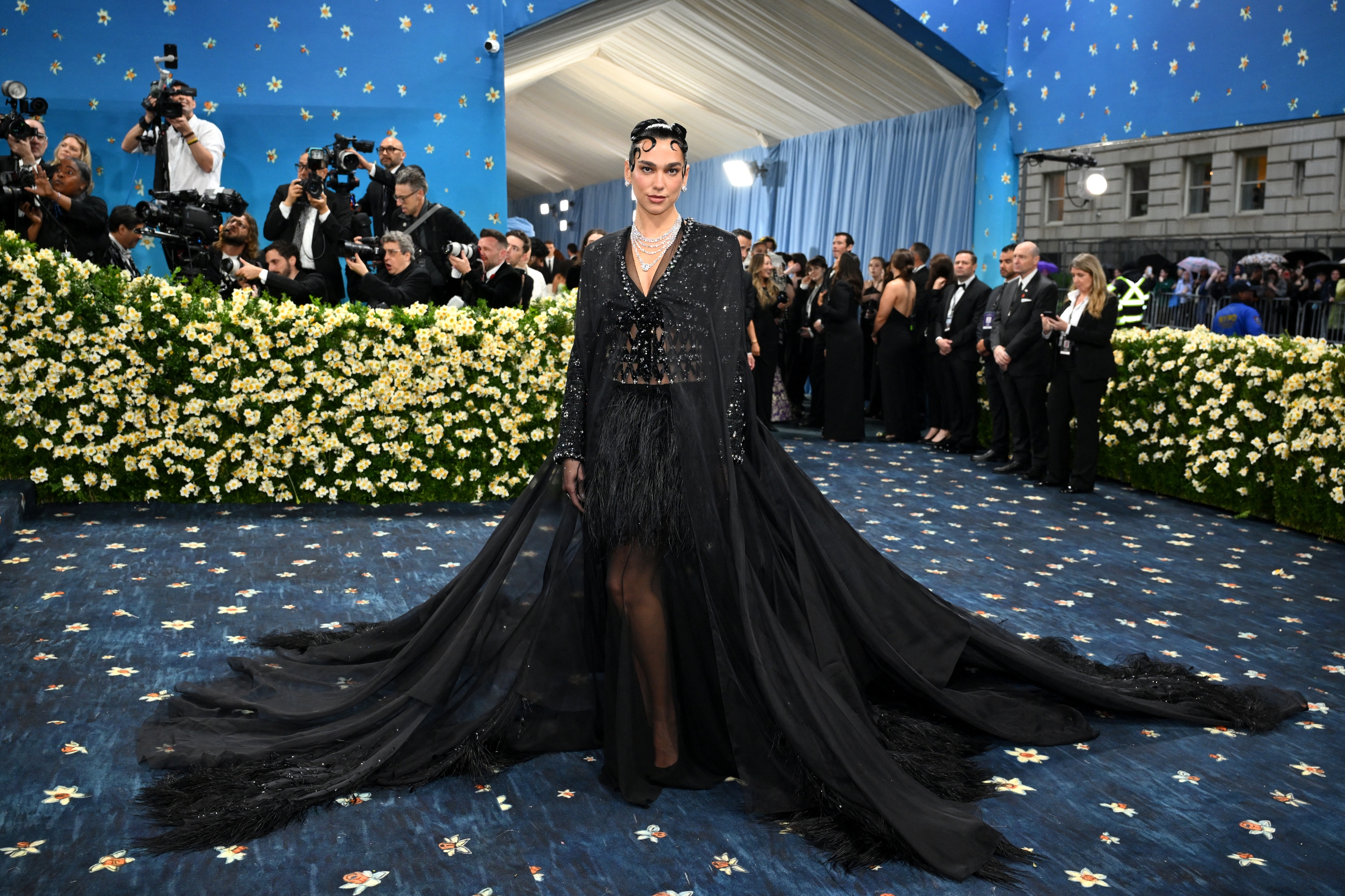 A person in an elegant, flowing black gown poses on a floral-themed red carpet with photographers in the background