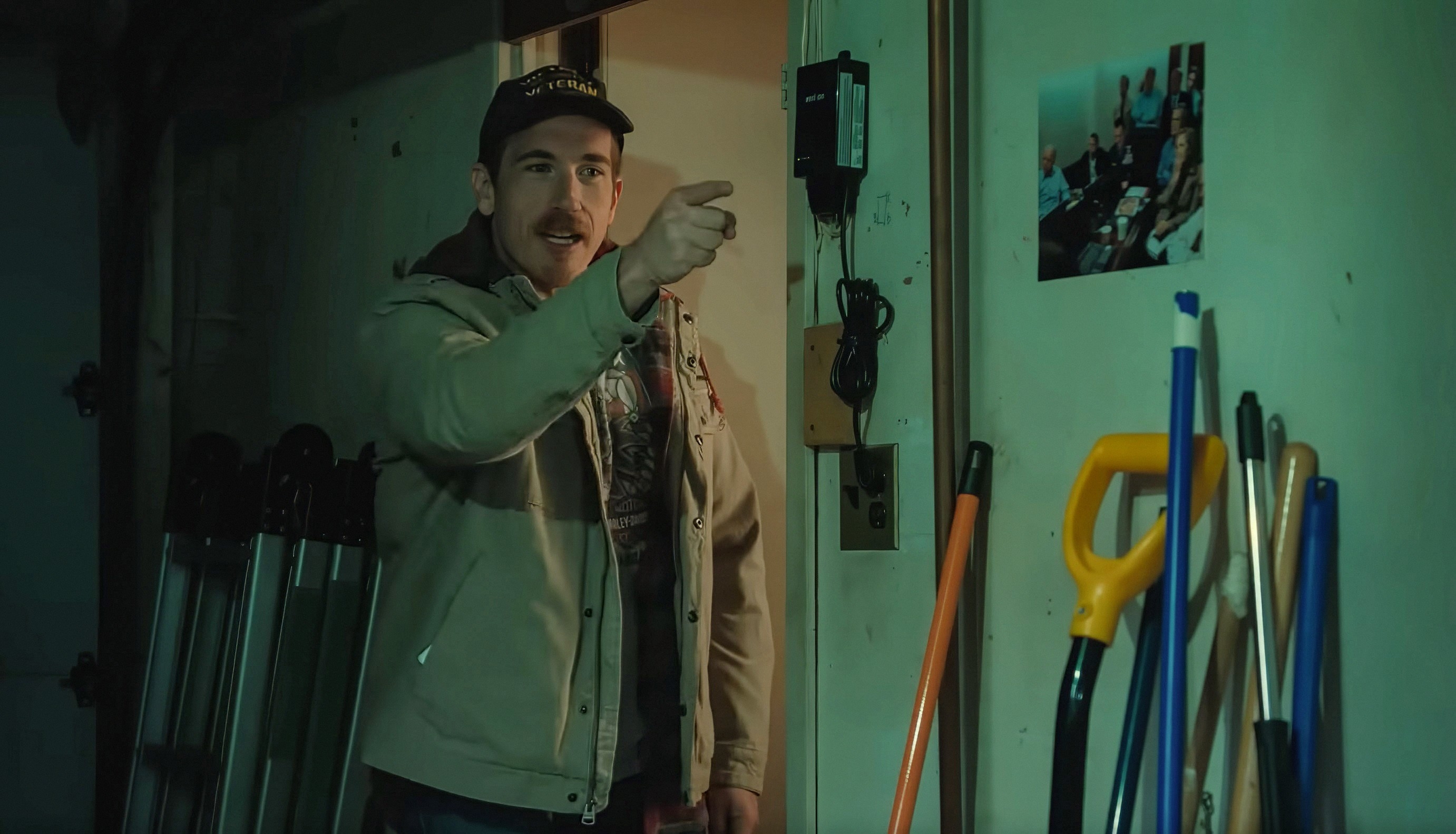 A person with a hat and jacket is pointing inside a garage or utility room, with various tools and a photo on the wall