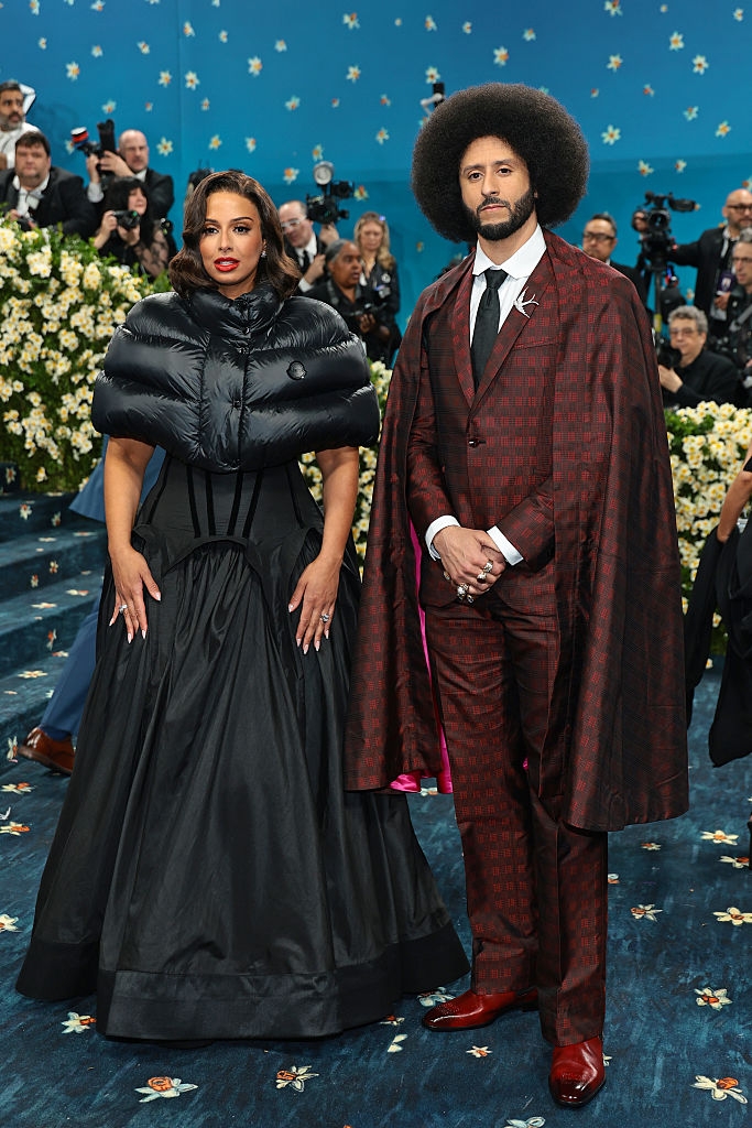 Nessa in a gown with puffy shoulders, Colin in a patterned suit with a long cape