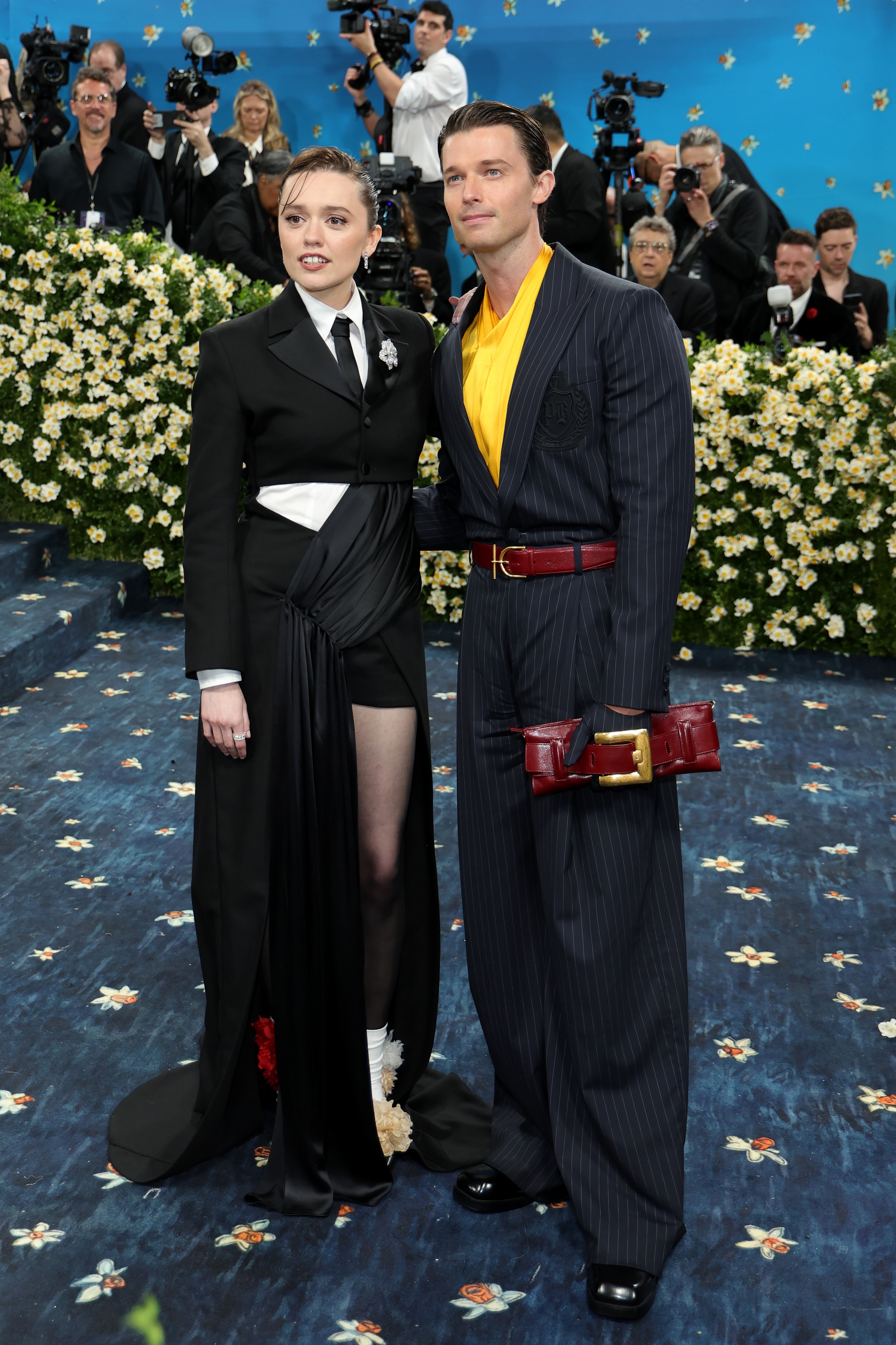 Two individuals on a red carpet: one in a black tuxedo with a skirt, the other in a pinstripe suit with a yellow shirt, posing for photographers