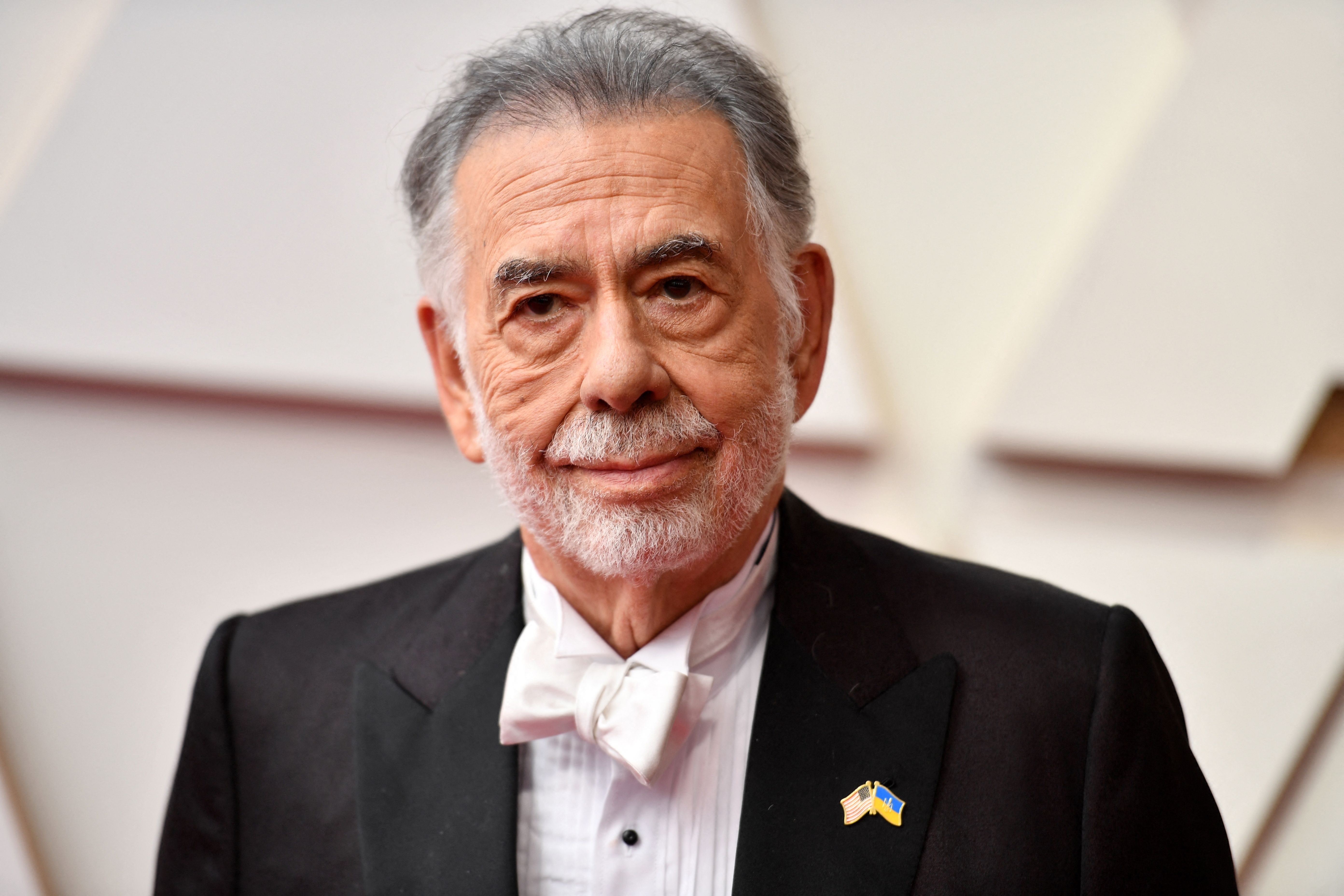 A distinguished older man in a tuxedo with a bow tie, standing on a red carpet at an event