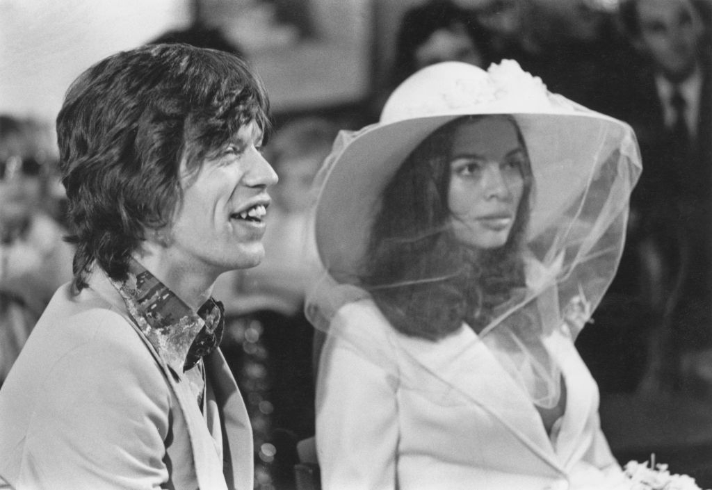 A man in a patterned shirt and suit sits beside a woman in a large hat with a veil. They appear to be attending an event