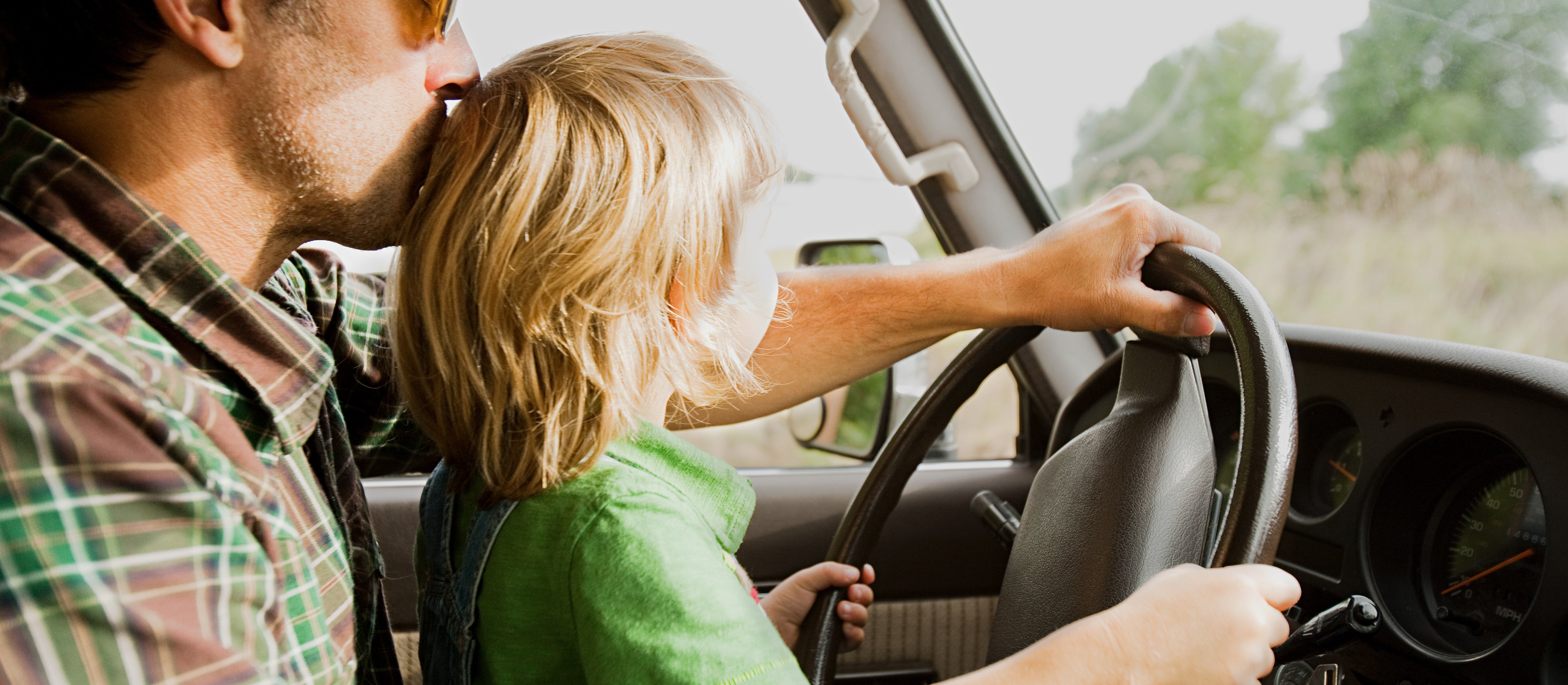 An adult kisses the head of a child sitting on their lap, playfully steering a car