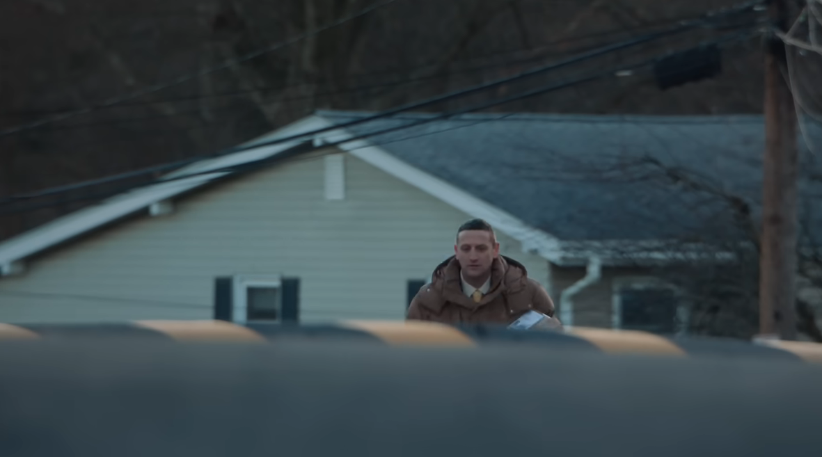 A person in a brown coat walks along a quiet street with houses in the background