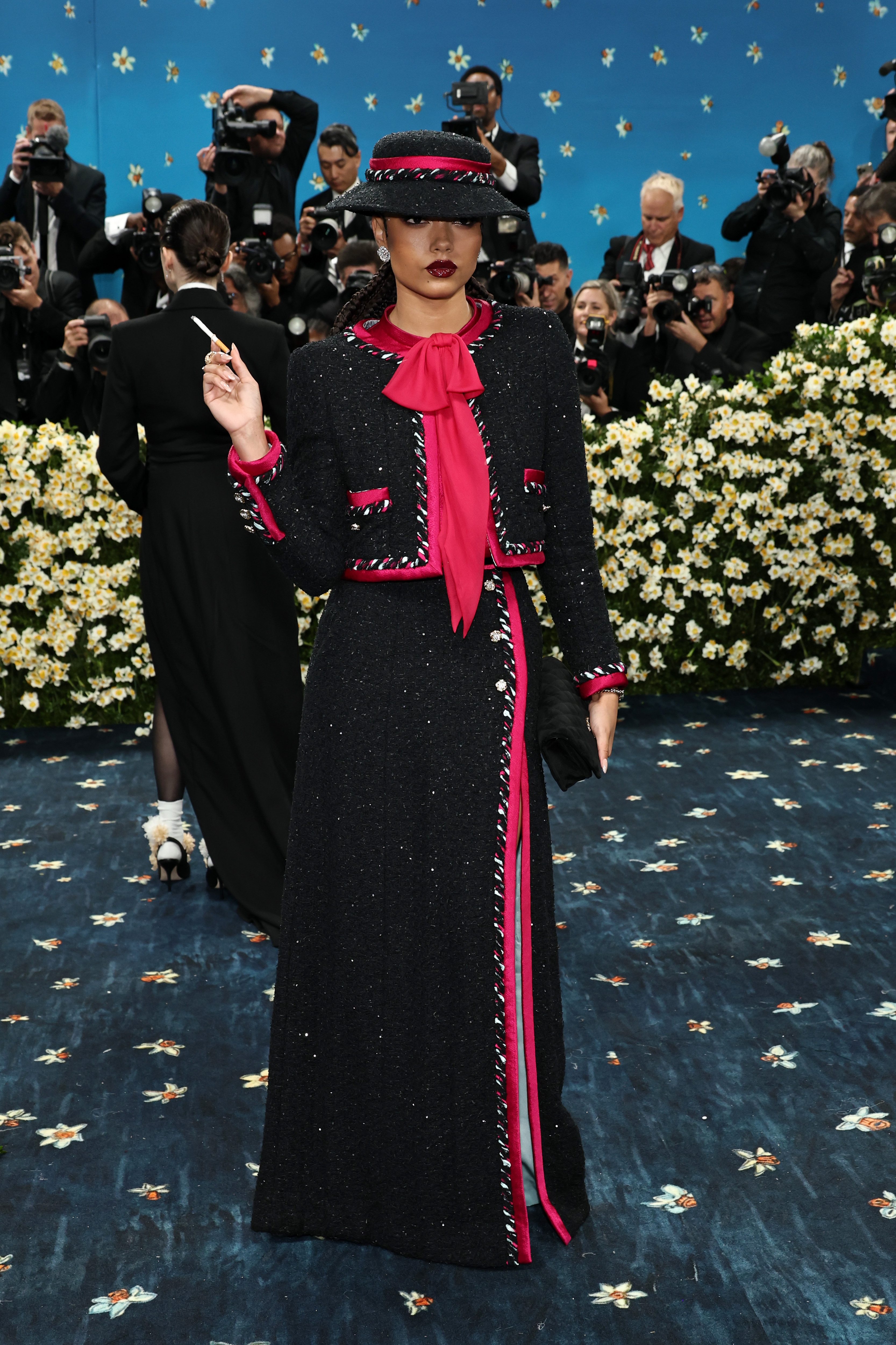 Person wearing a vintage-inspired black tweed suit with pink accents and wide-brimmed hat on a red carpet, surrounded by photographers