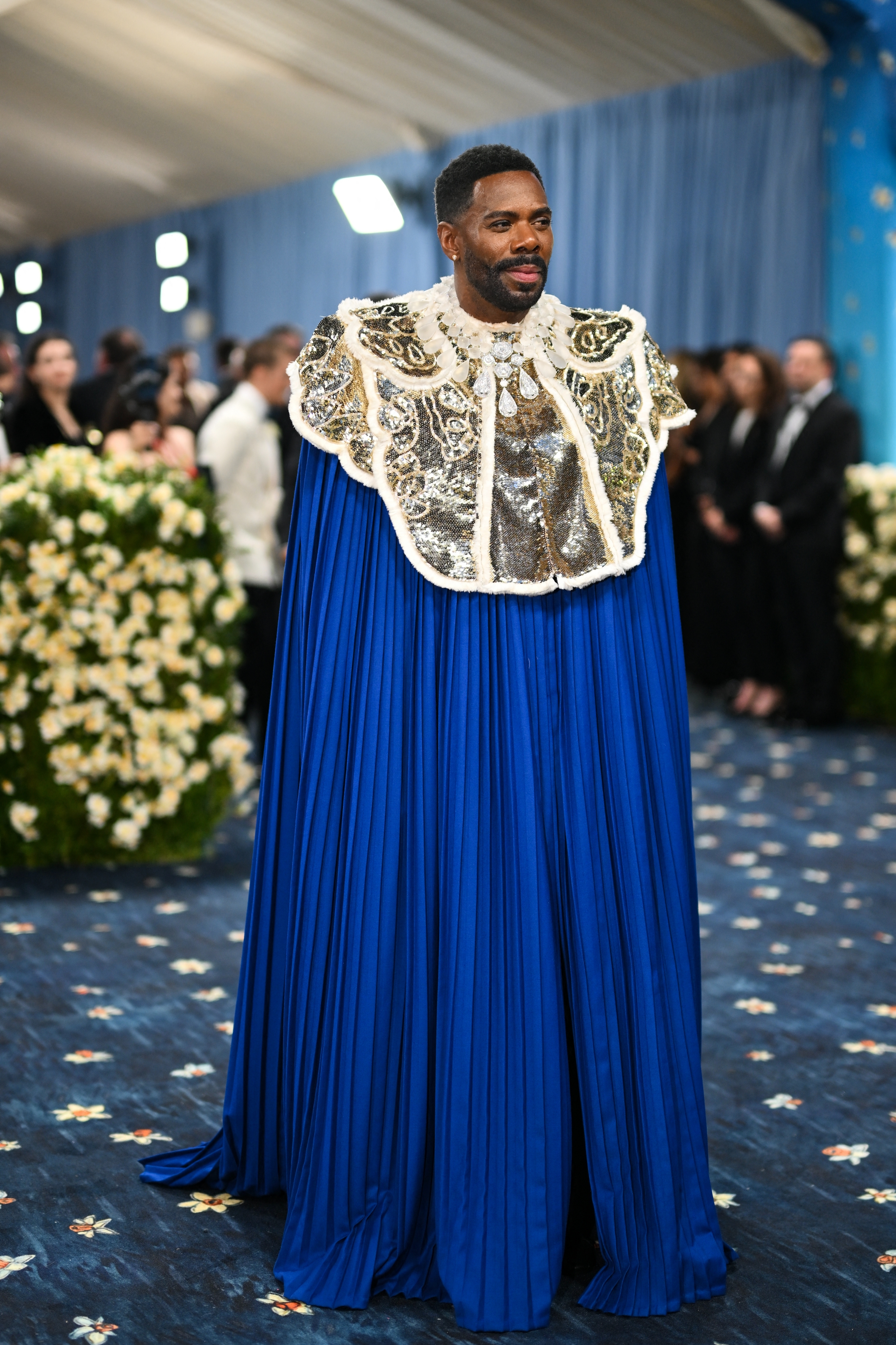 Colman Domingo on the red carpet wearing an ornate cape with intricate patterns and pleated fabric, attended by event guests in the background