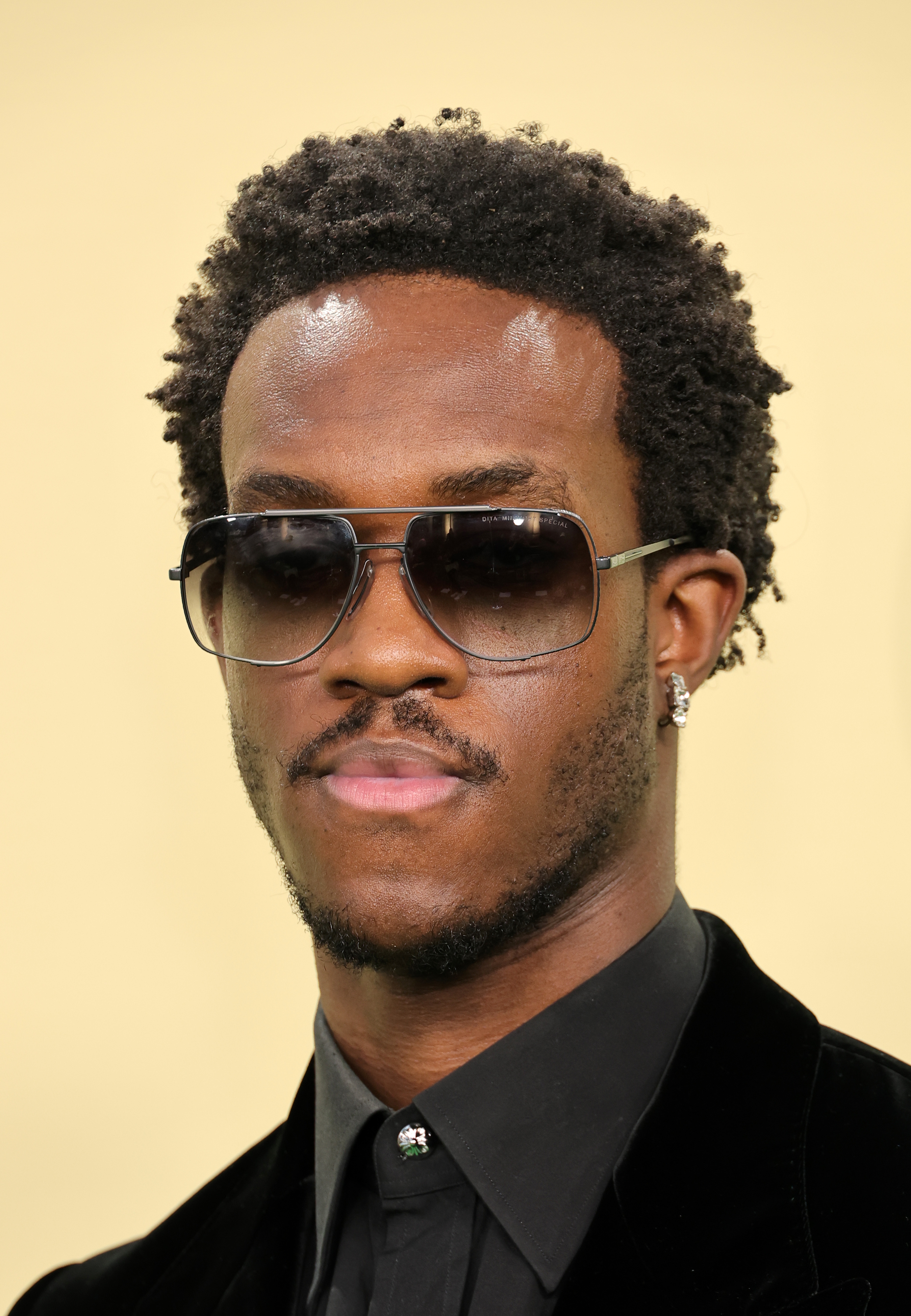 Wisdom Kaye wearing sunglasses and a black suit with a collared shirt, looking serious