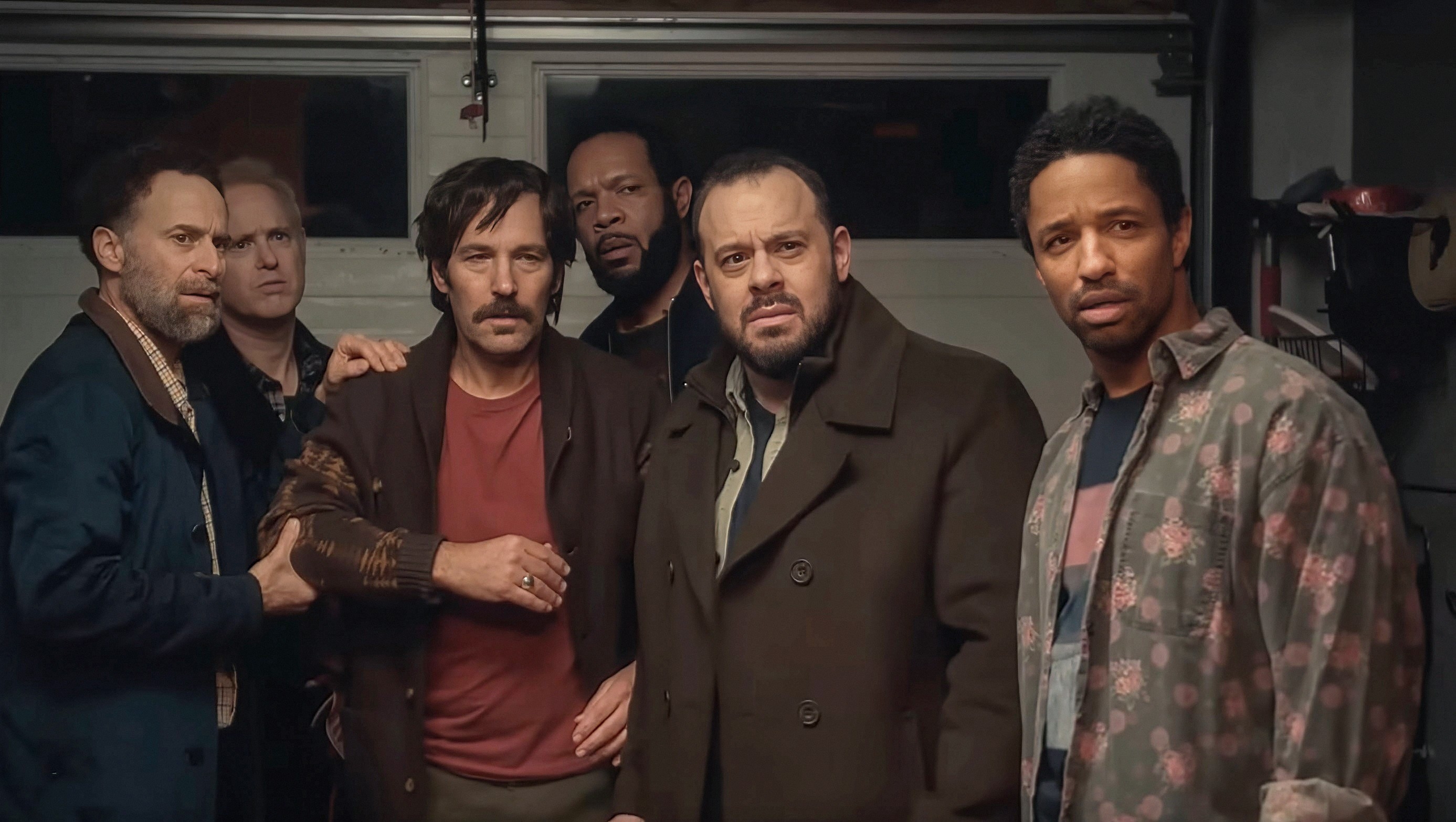 Six men stand in a garage, looking intently at the camera. They appear serious, engaged in a tense or dramatic moment
