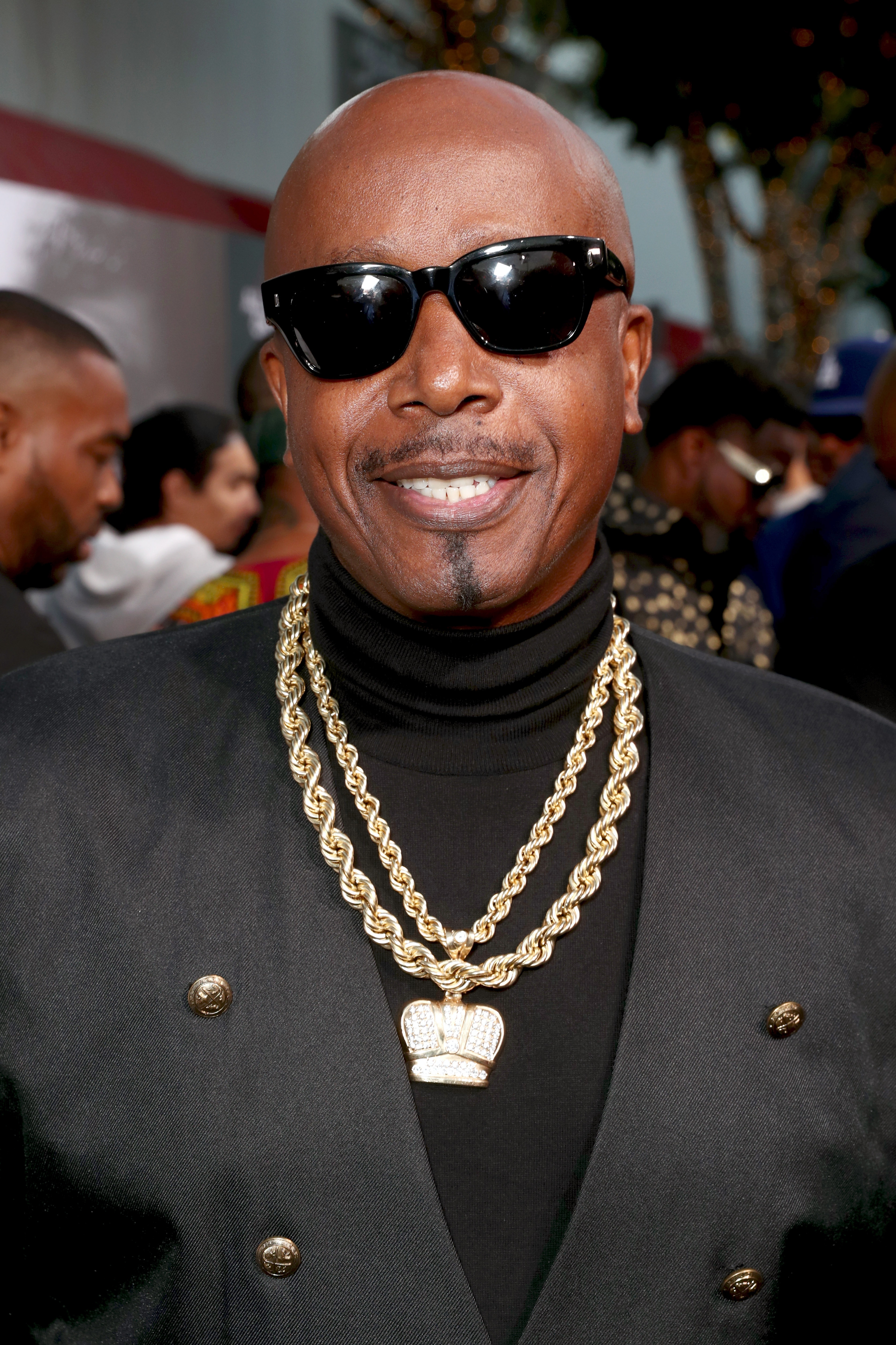 A person wearing sunglasses and a stylish black suit with gold chains smiles at a red carpet event
