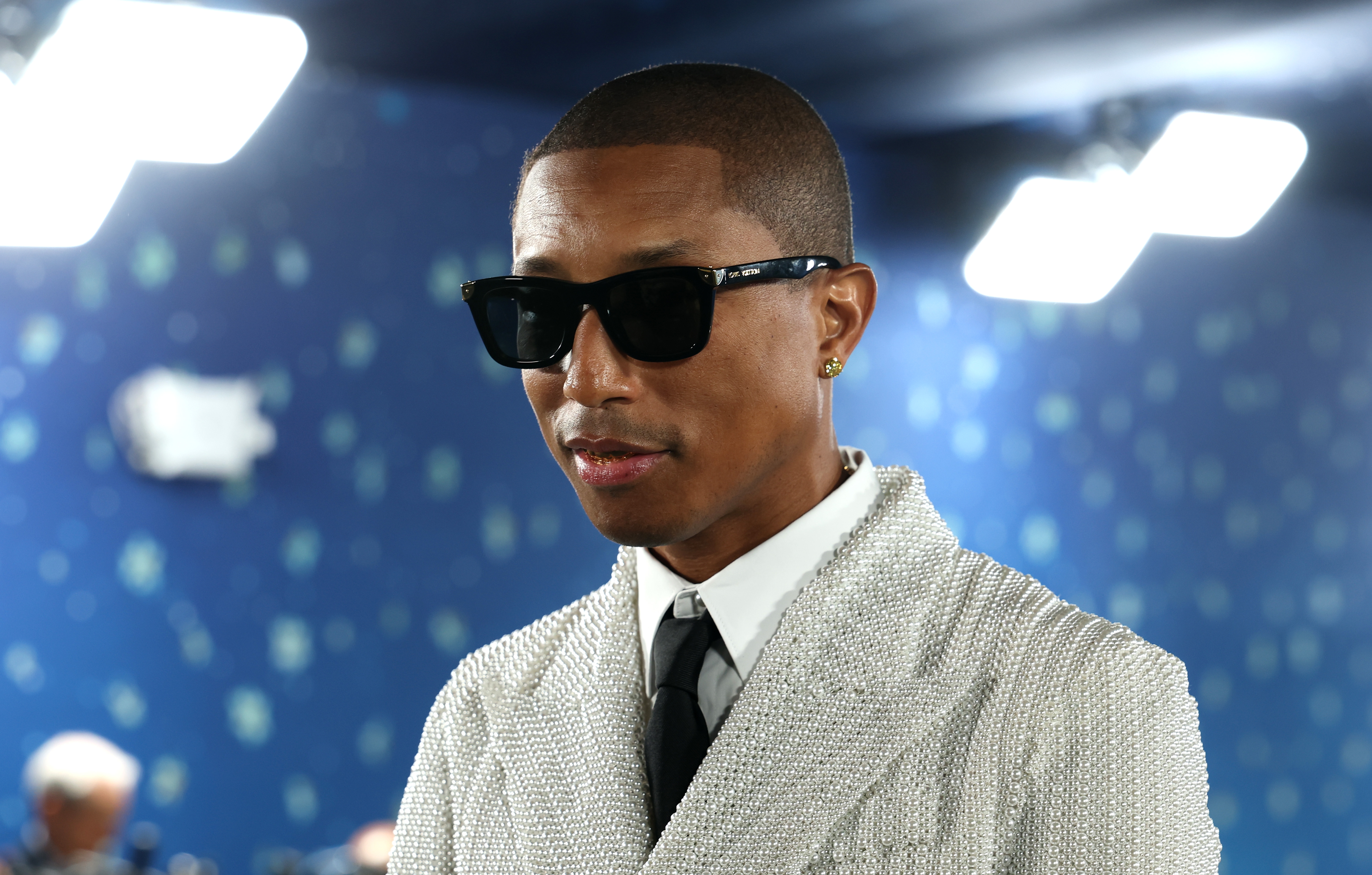 Pharrell in a textured suit and tie, wearing dark sunglasses, standing indoors with a patterned starry background