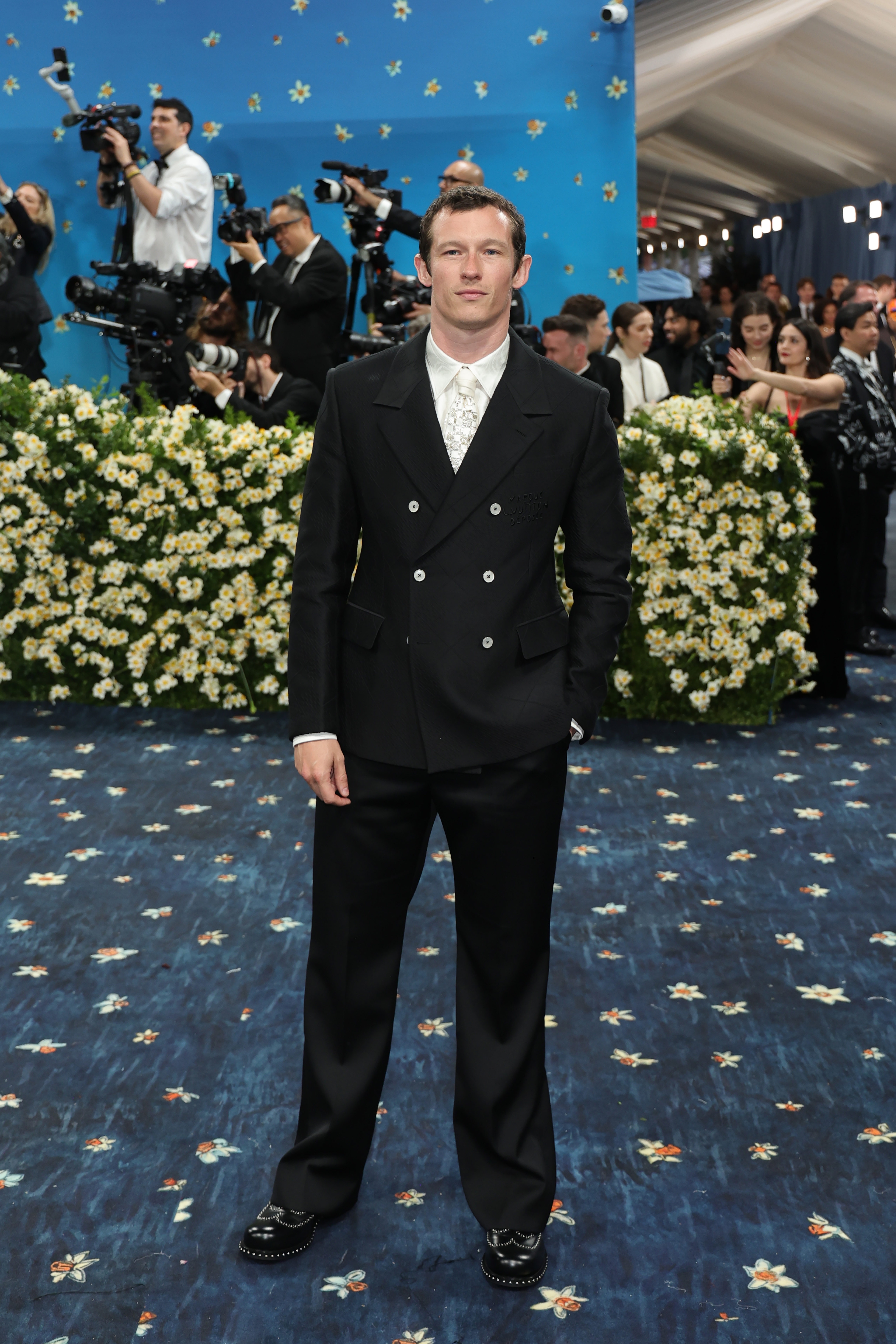A person in a double-breasted suit poses on a floral-patterned carpet at a high-profile event, with photographers capturing the moment