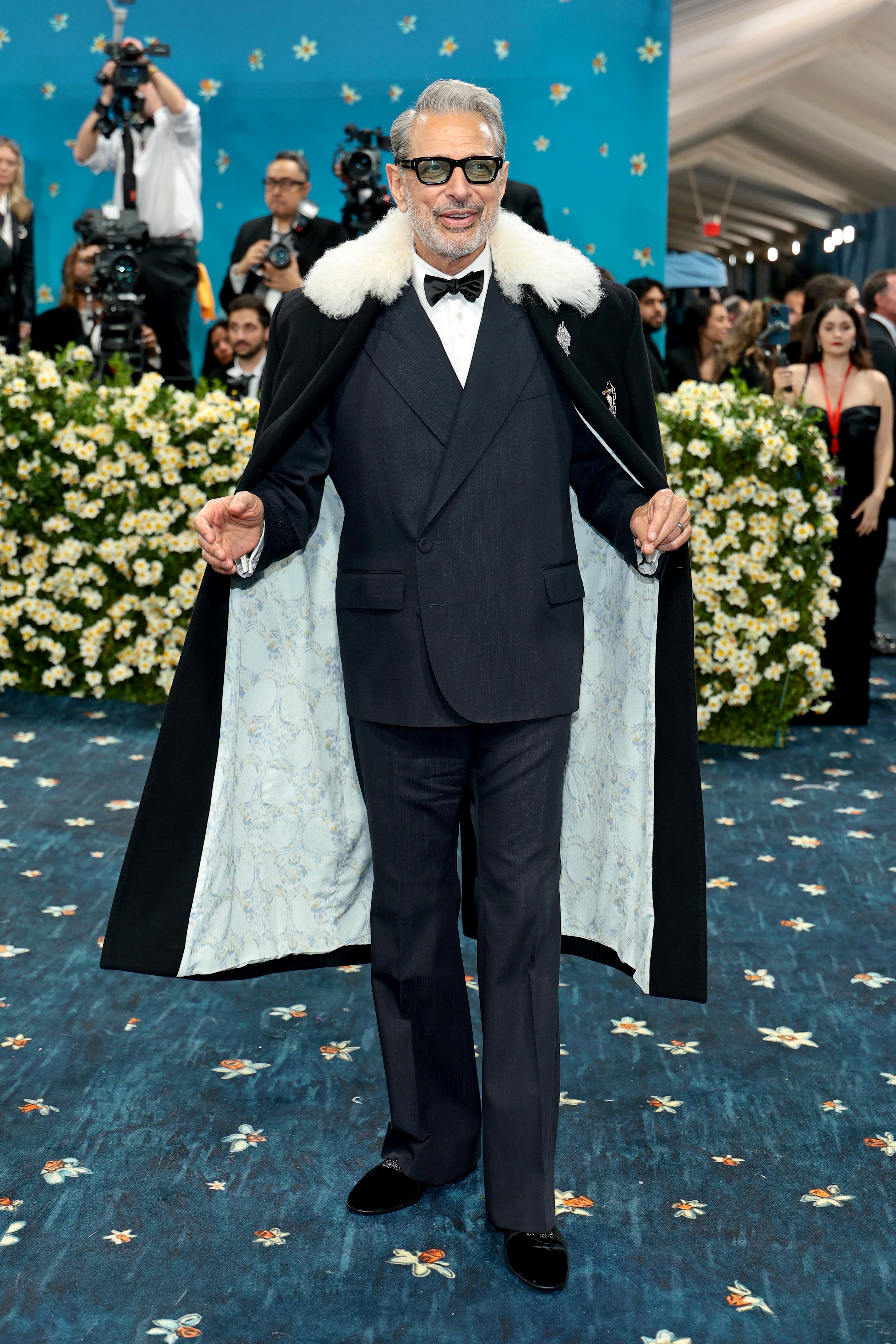 Man in a classic tuxedo with glasses and a white fur-collared coat poses on a floral-patterned carpet at a formal celebrity event