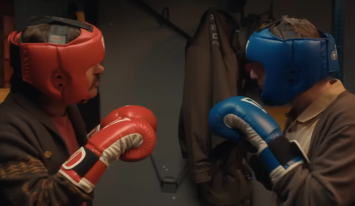Two people face each other wearing protective headgear and boxing gloves in a sparring stance inside a gym or training room