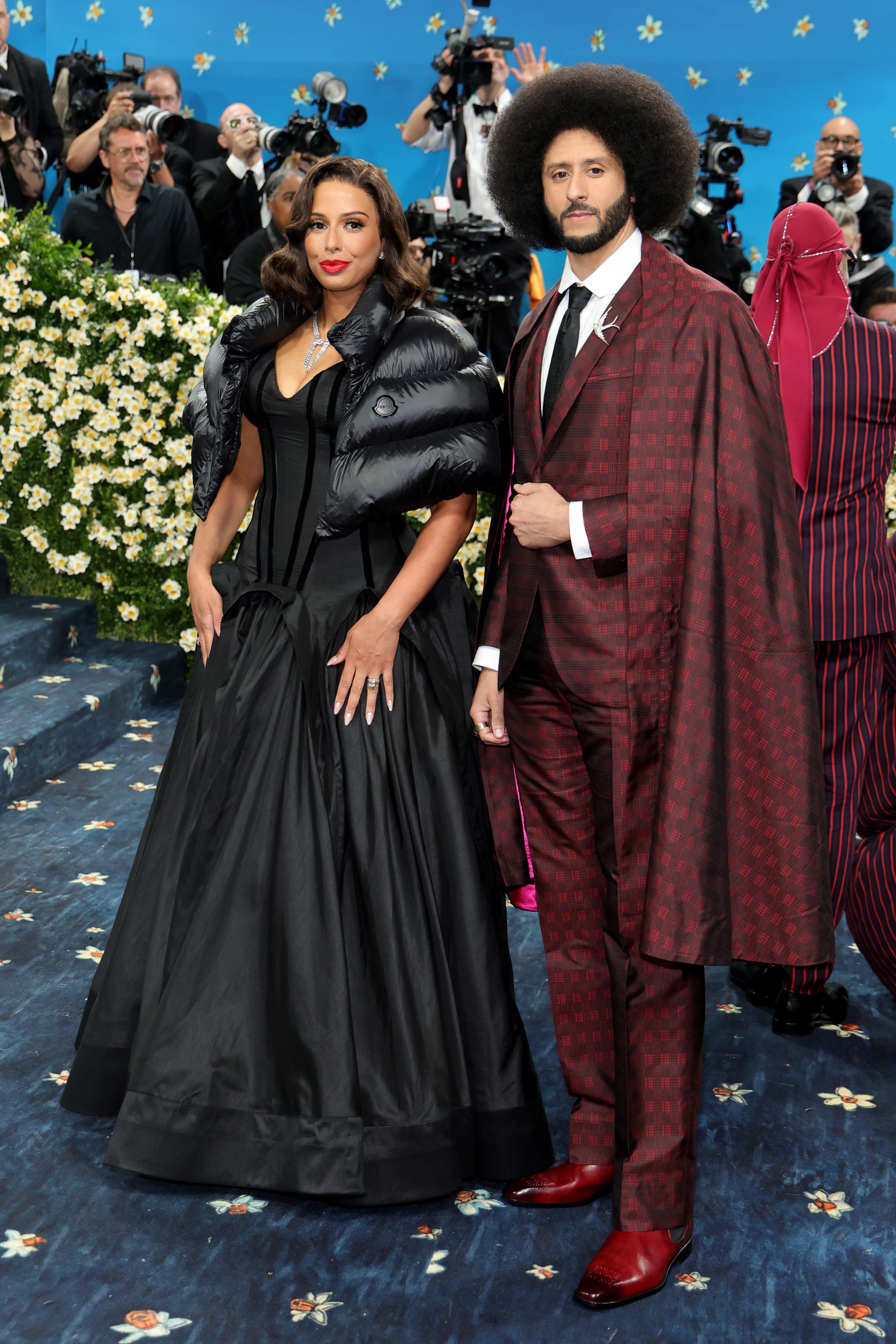 Two people on a blue carpet event. The person on the left wears a black gown with a puffy shoulder wrap, the person on the right wears a patterned burgundy suit with a matching cape