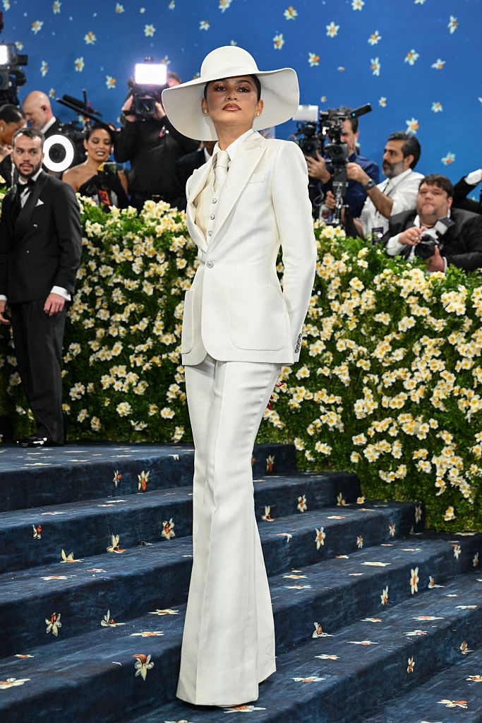 Person in an elegant tailored suit and wide-brimmed hat poses on a floral-themed red carpet with photographers in the background