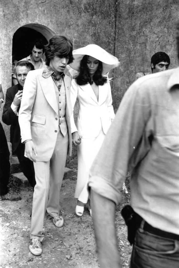 A couple in stylish vintage suits and hats walks hand in hand, surrounded by onlookers, evoking a classic, elegant vibe