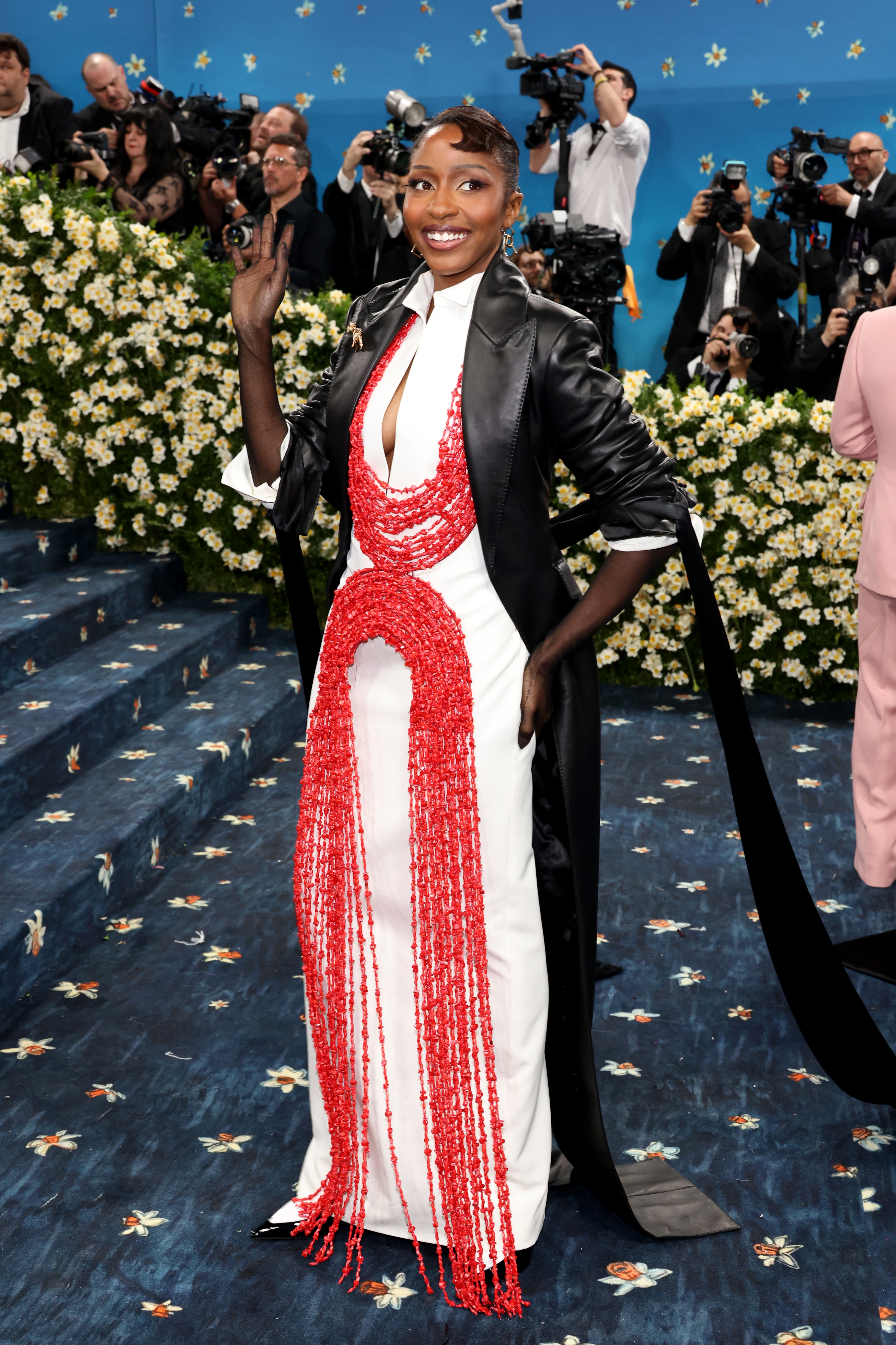 Person in a white gown with red beaded detailing and a black coat on a blue carpet, waving to photographers