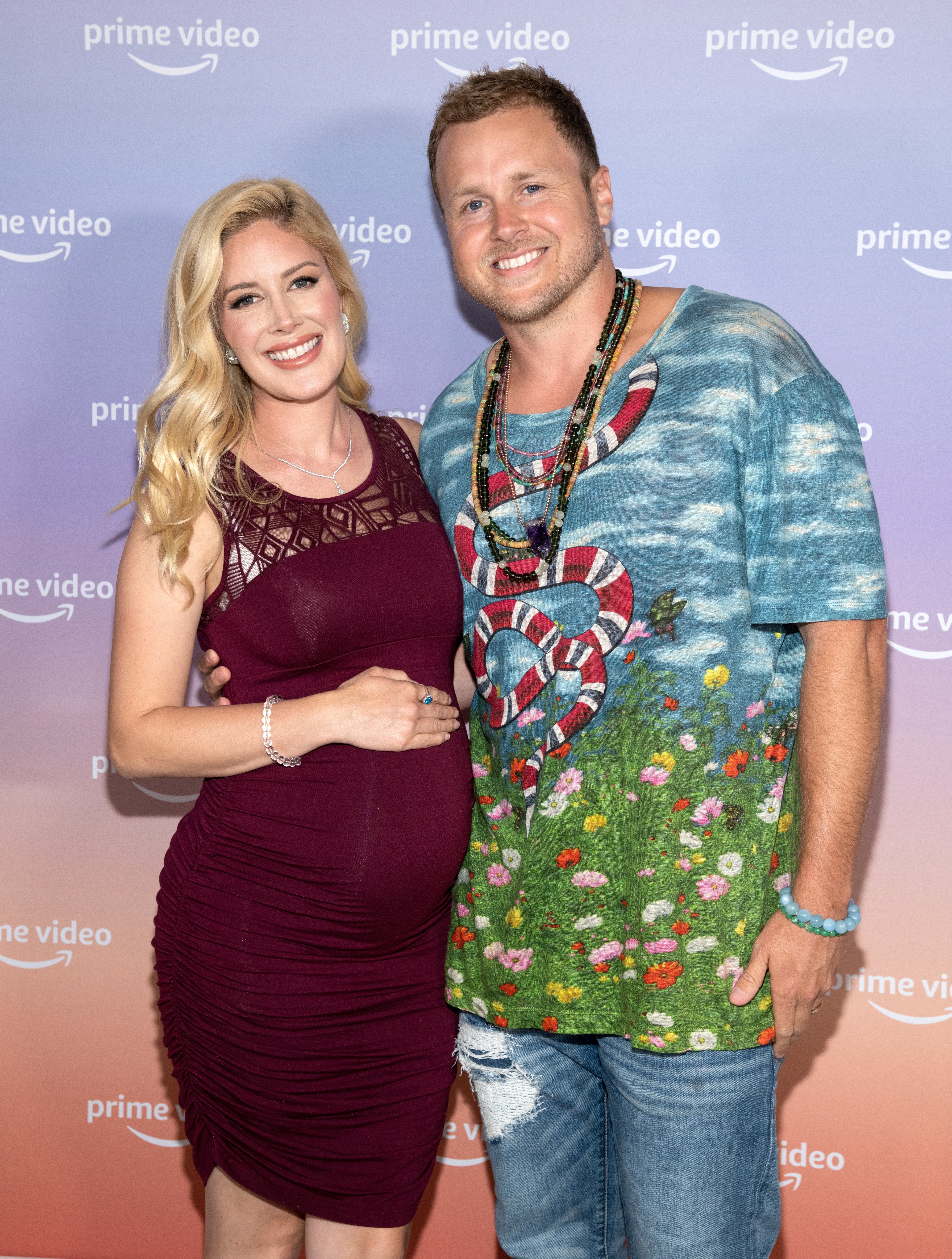 A smiling pregnant woman in a fitted lace dress and a man in a printed t-shirt and jeans, posing together at a Prime Video event
