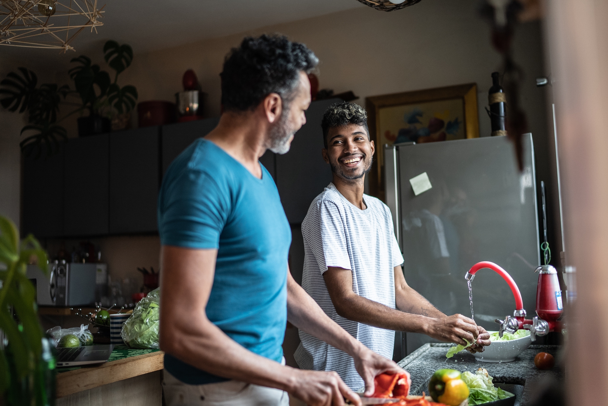Two smiling men preparing food in a kitchen, one washing veggies, the other chopping. They appear engaged and happy together