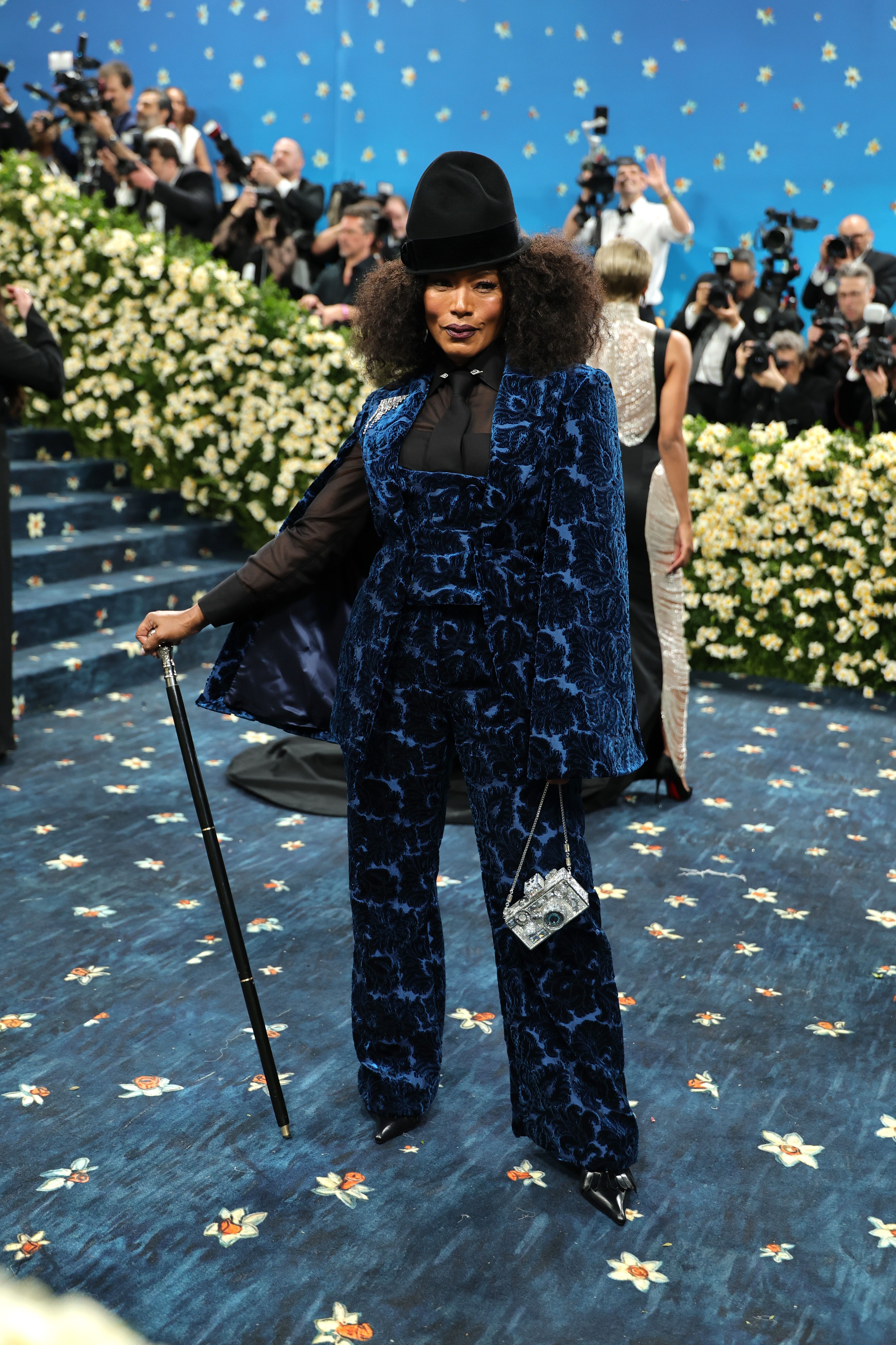 Person in a stylish, patterned pantsuit and wide-brimmed hat on a floral carpet, holding a cane, at a celebrity event