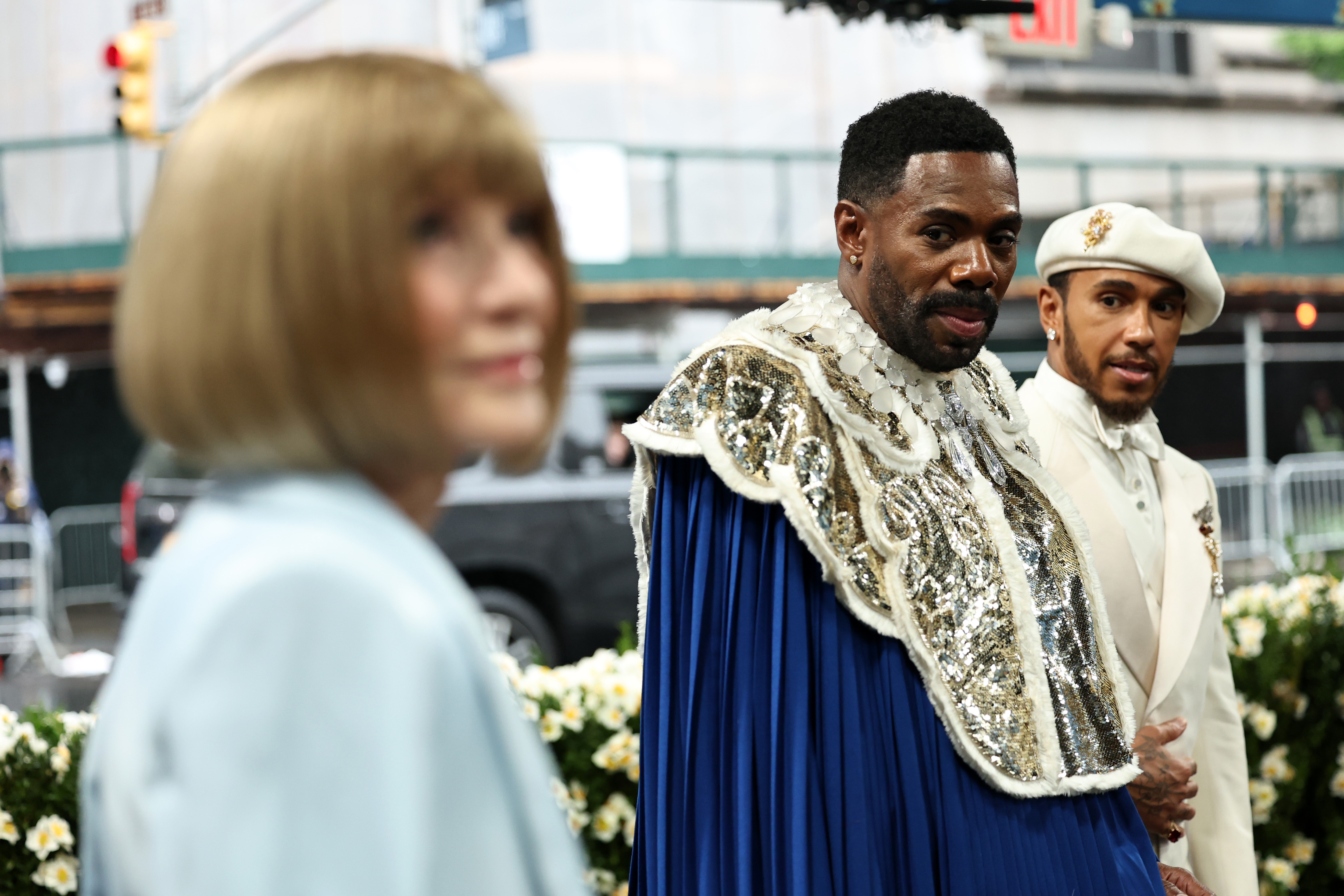 Three individuals on a red carpet; one wears a bejeweled cape, another in a cream outfit and cap, and a third with a bob haircut in profile