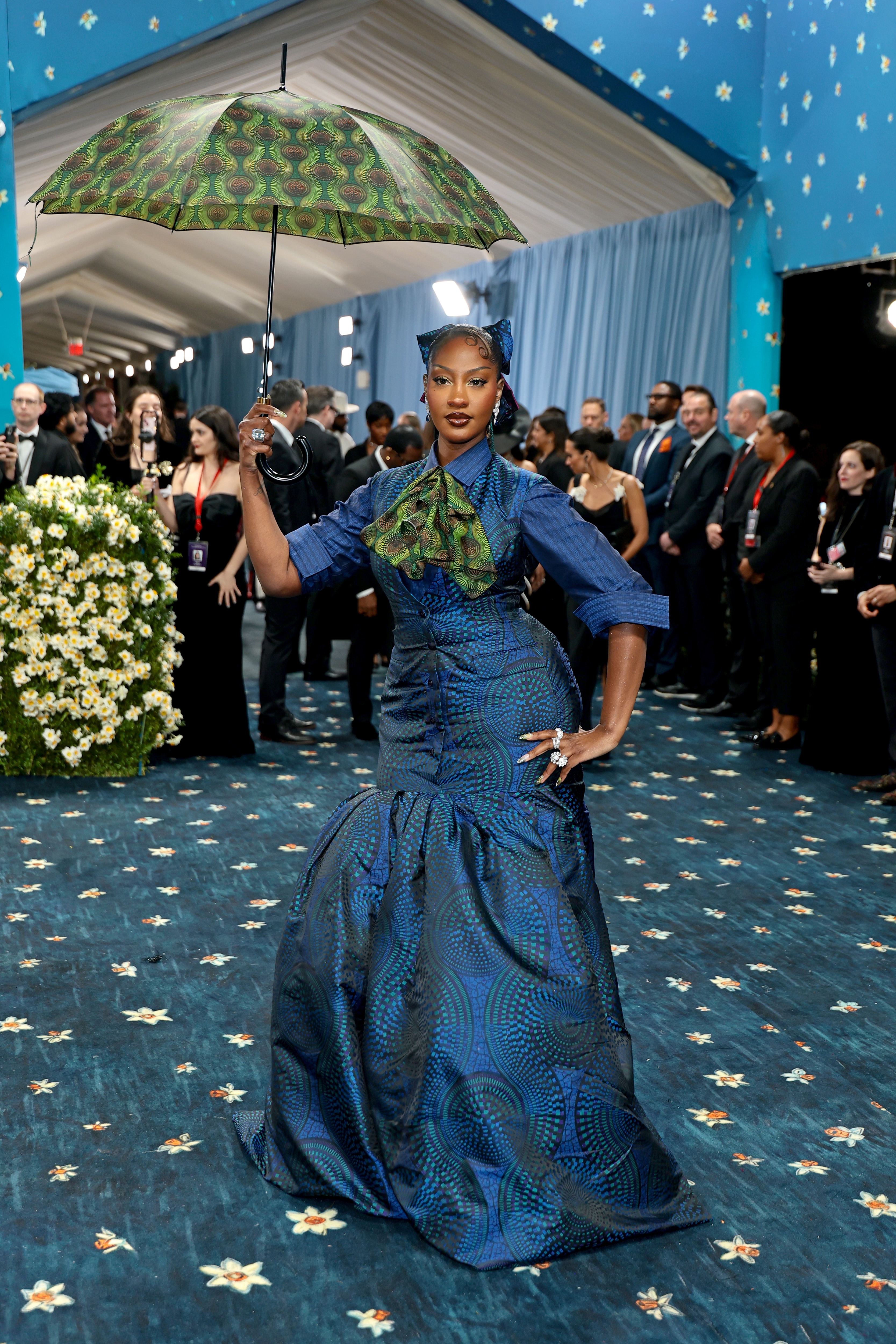 A person poses on a blue-themed carpet in a patterned gown with matching umbrella and headpiece, surrounded by event attendees
