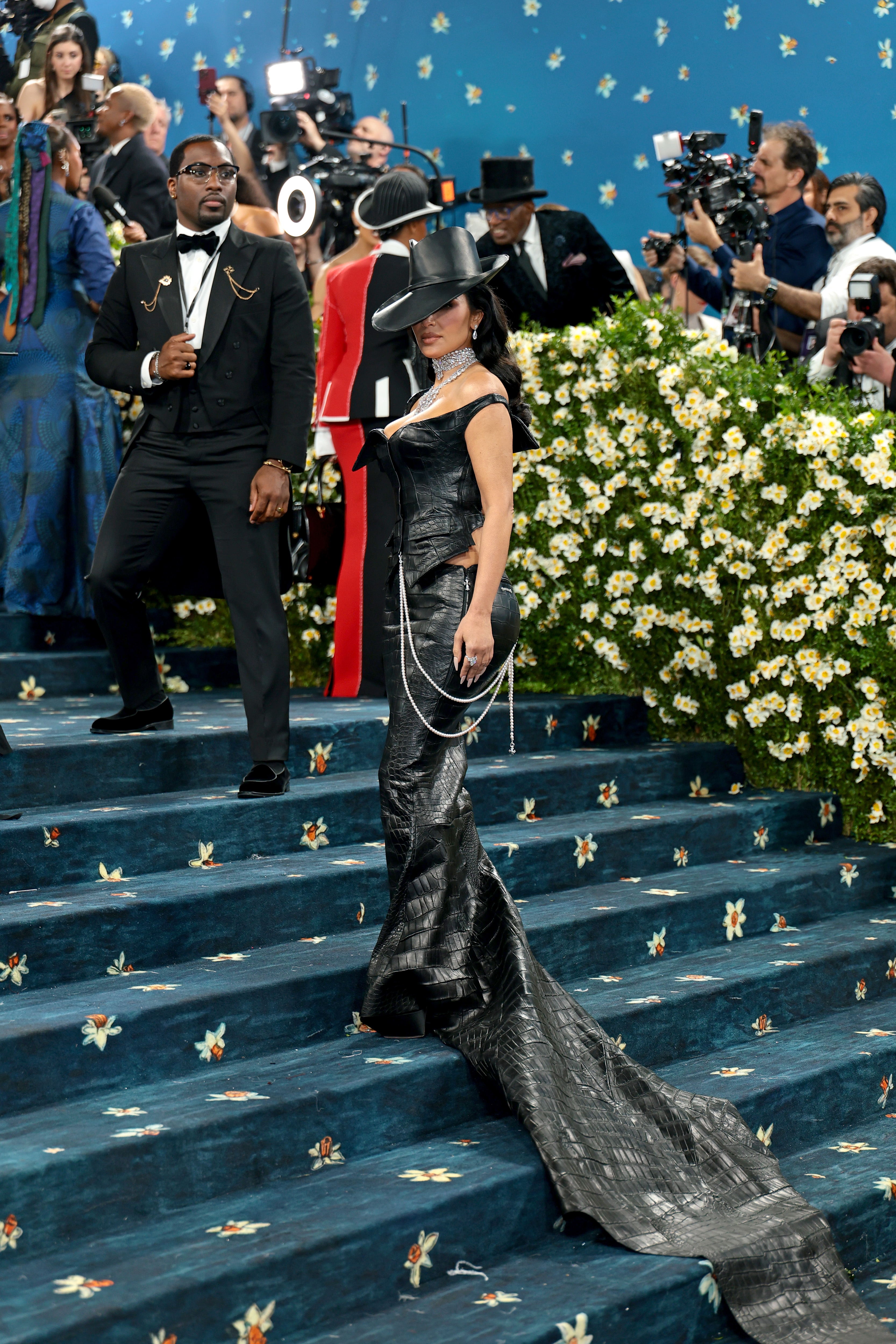 Two celebrities on a floral-decorated staircase, one in a black tuxedo, the other in a dramatic black dress with a long train and wide-brimmed hat
