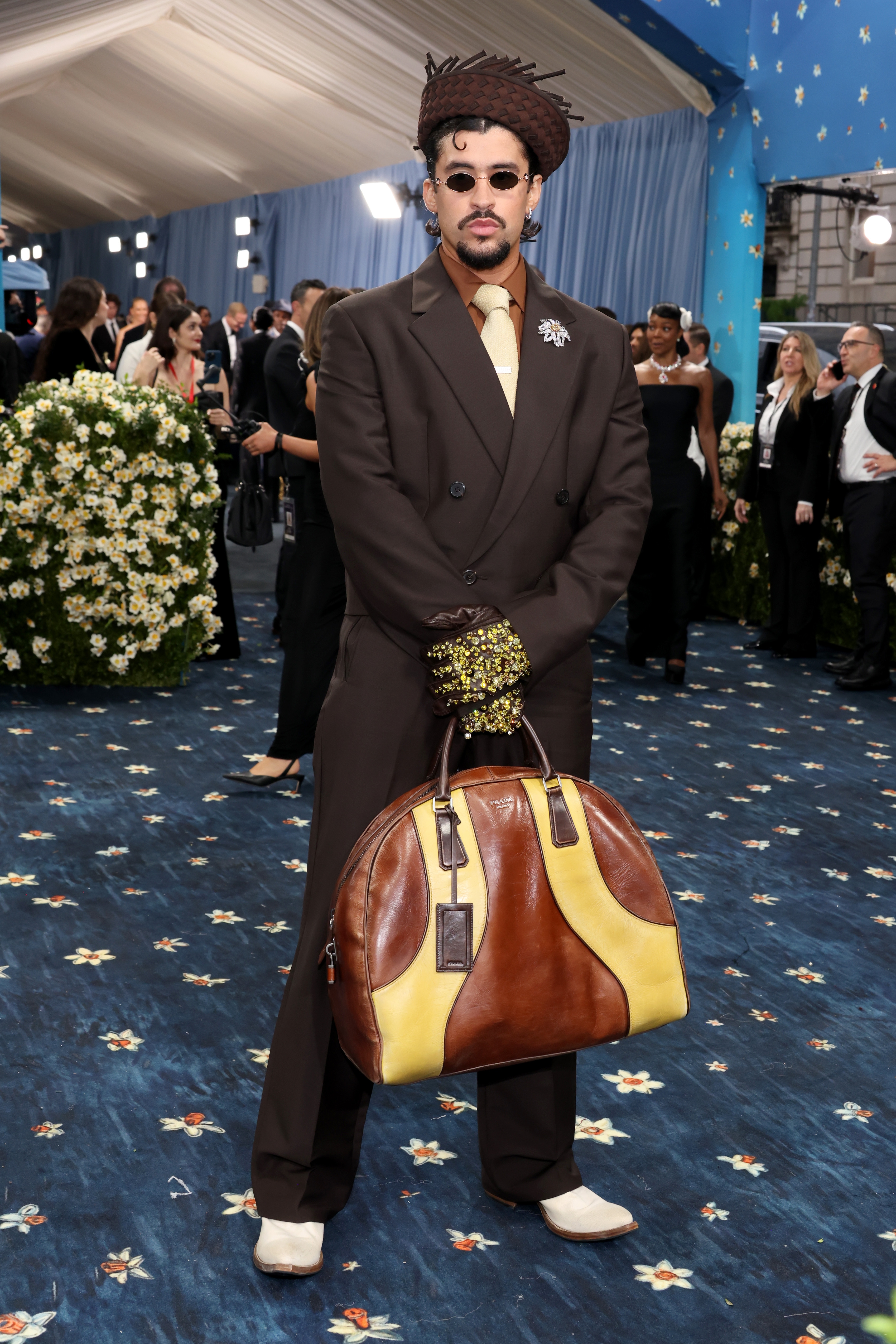 Person in unique braided hat and brown suit with floral brooch, patterned gloves, and large brown-and-yellow handbag, standing on floral carpet