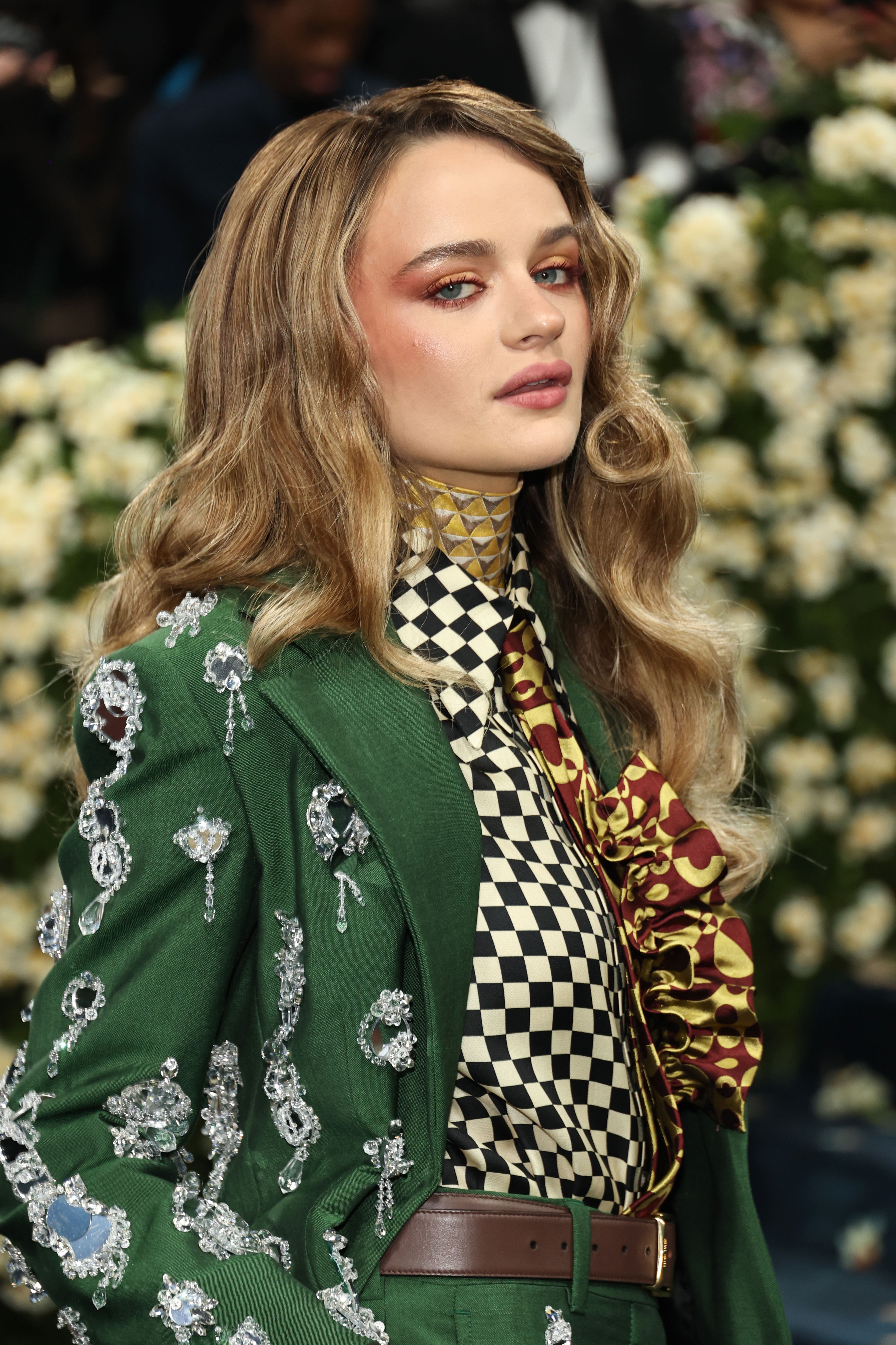 Person on red carpet wearing a green suit with floral embellishments, checkered shirt, and patterned scarf, in front of a floral backdrop