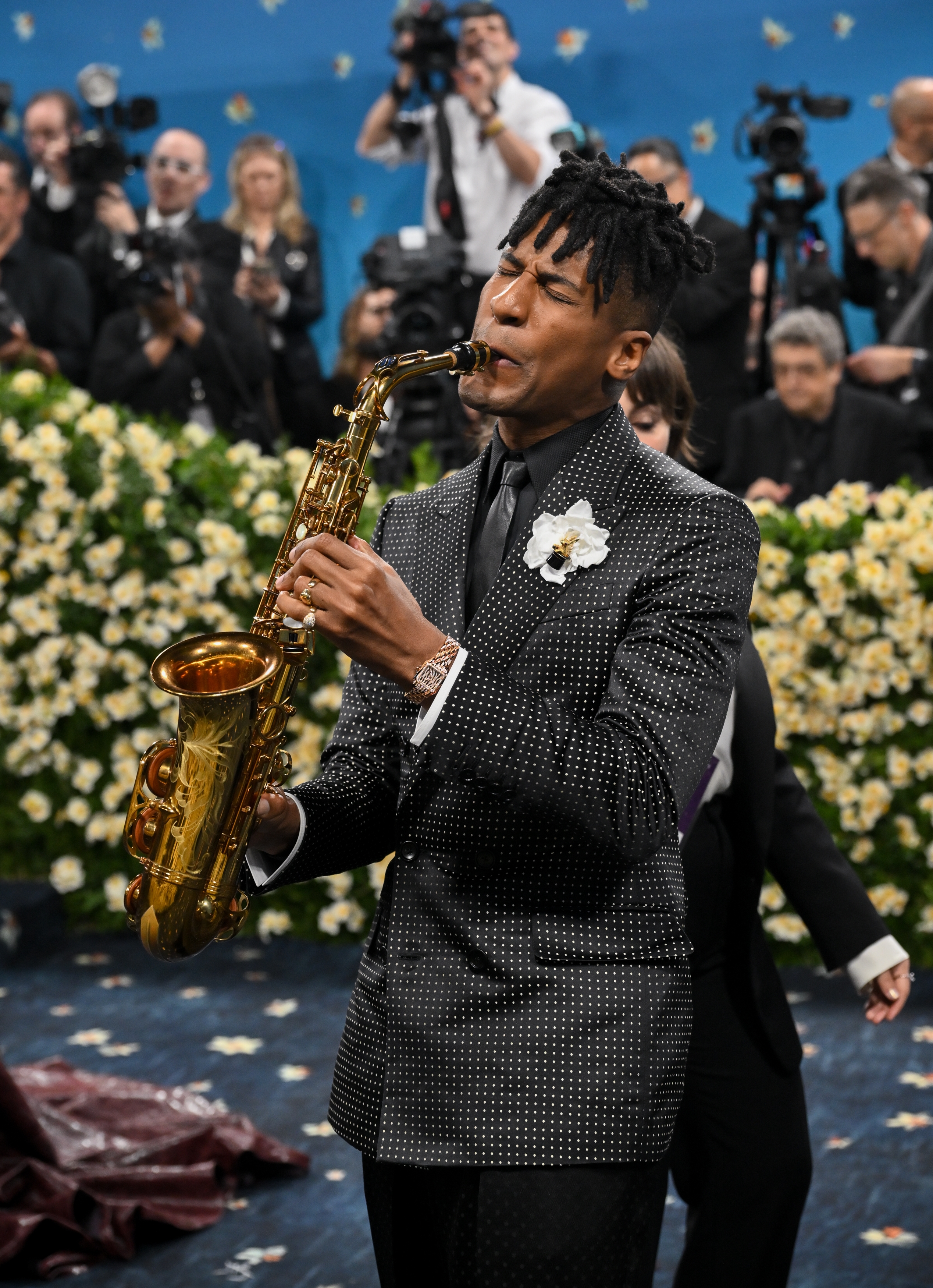 Jon Batiste playing saxophone on the red carpet, wearing a polka dot-patterned suit with floral boutonnière, surrounded by photographers