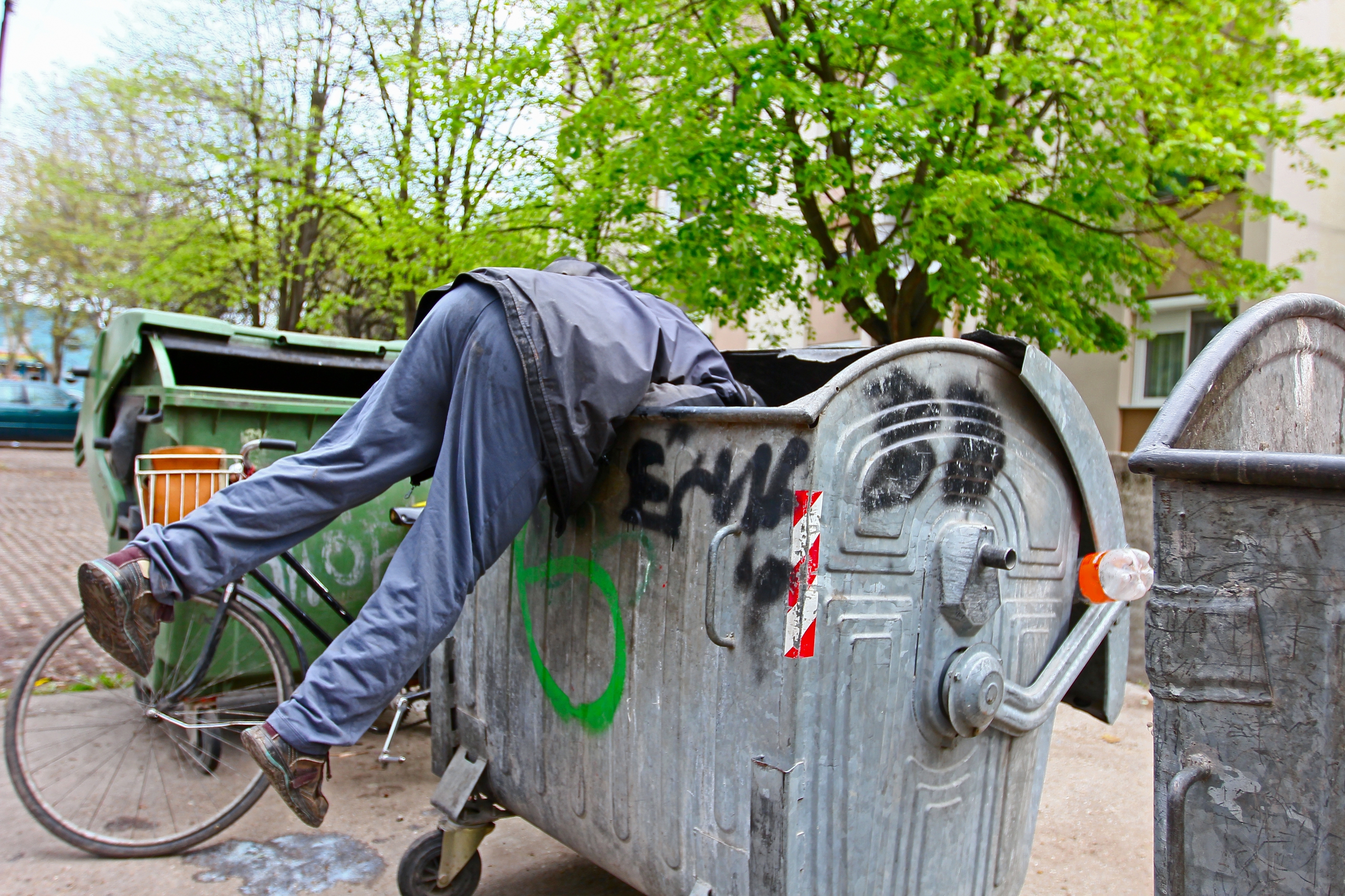 Person in casual clothes leaning into a large metal dumpster with legs sticking out, next to a parked bicycle on a street