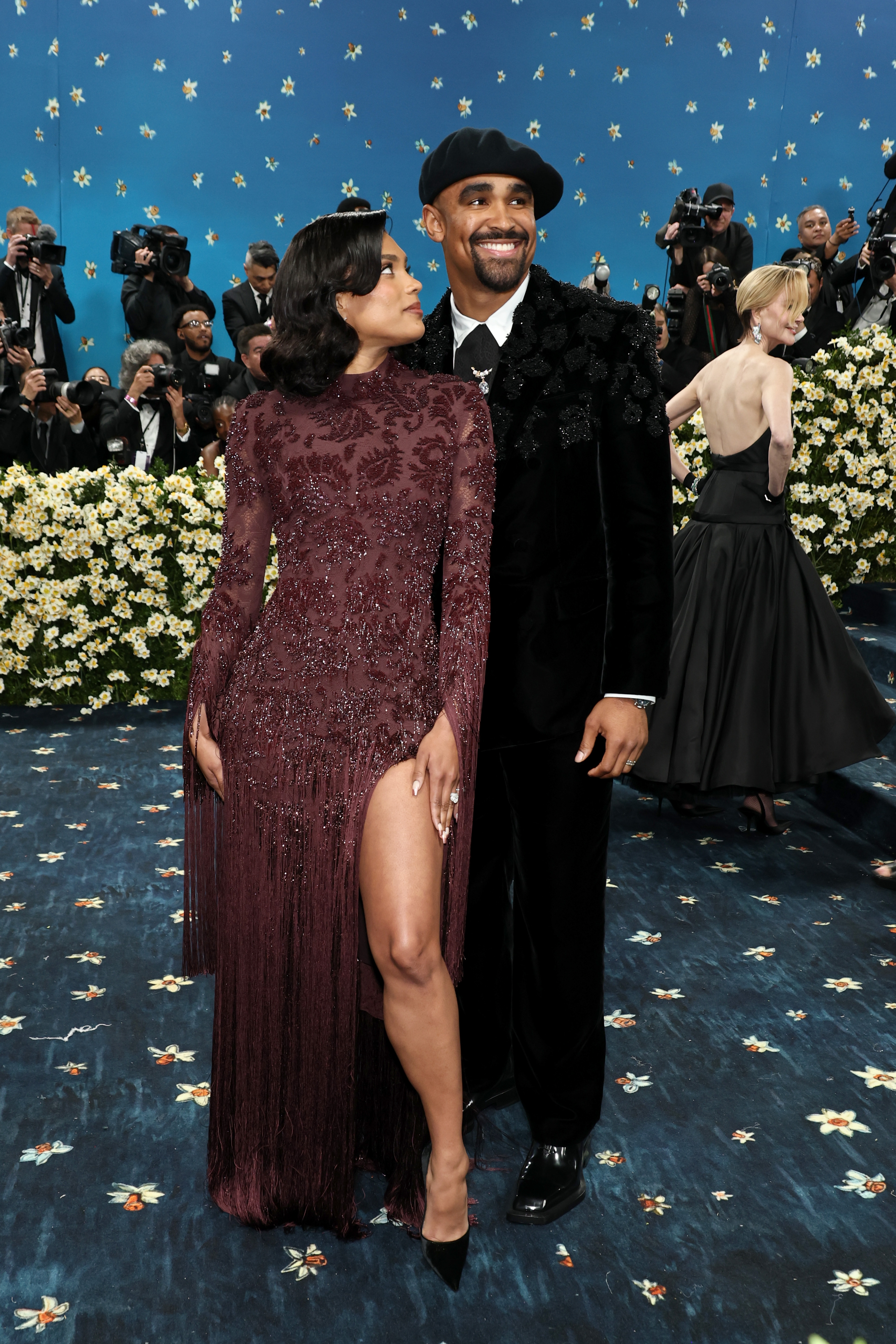 Two individuals on a red carpet; one wearing a long, elegant dress with a high slit, the other in a stylish suit. Photographers are in the background