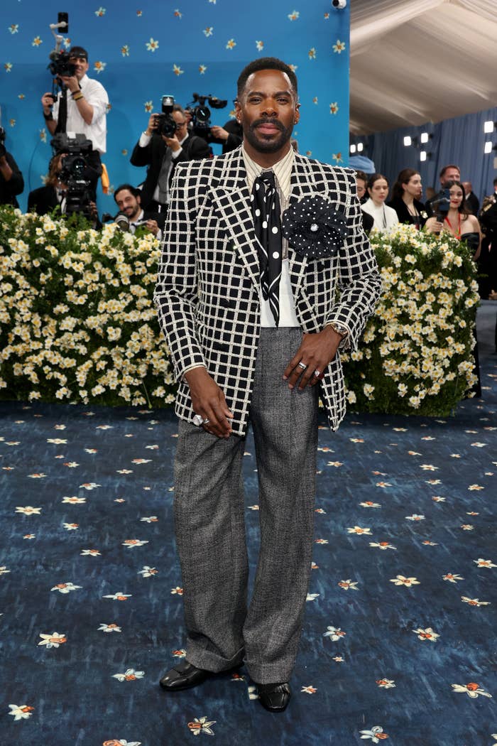 Person in stylish checkered suit and wide-leg pants on a floral-themed red carpet, posing confidently while photographers capture the moment