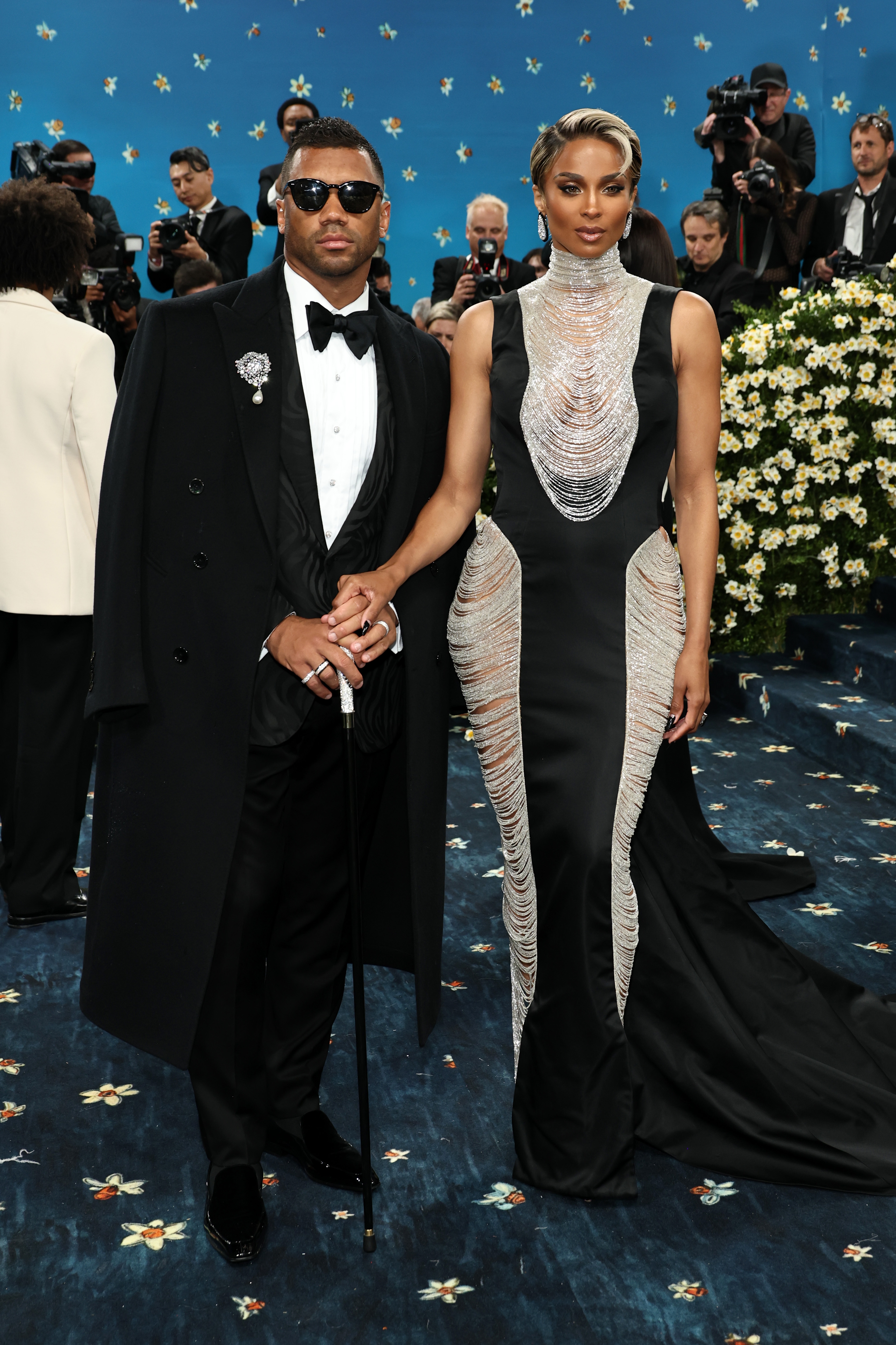 Two celebrities on a red carpet; the man in a tuxedo with sunglasses, the woman in a form-fitting gown with intricate silver detailing