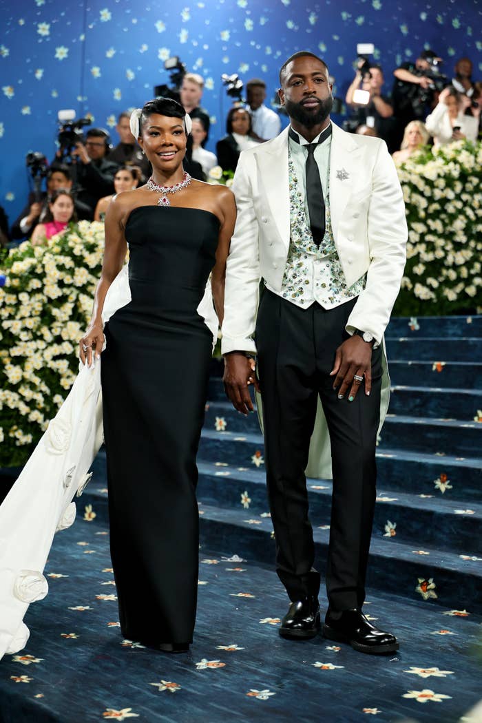 Two individuals on a floral-themed event stairway. Woman in elegant strapless gown with train; man in black tuxedo with floral vest and white coat