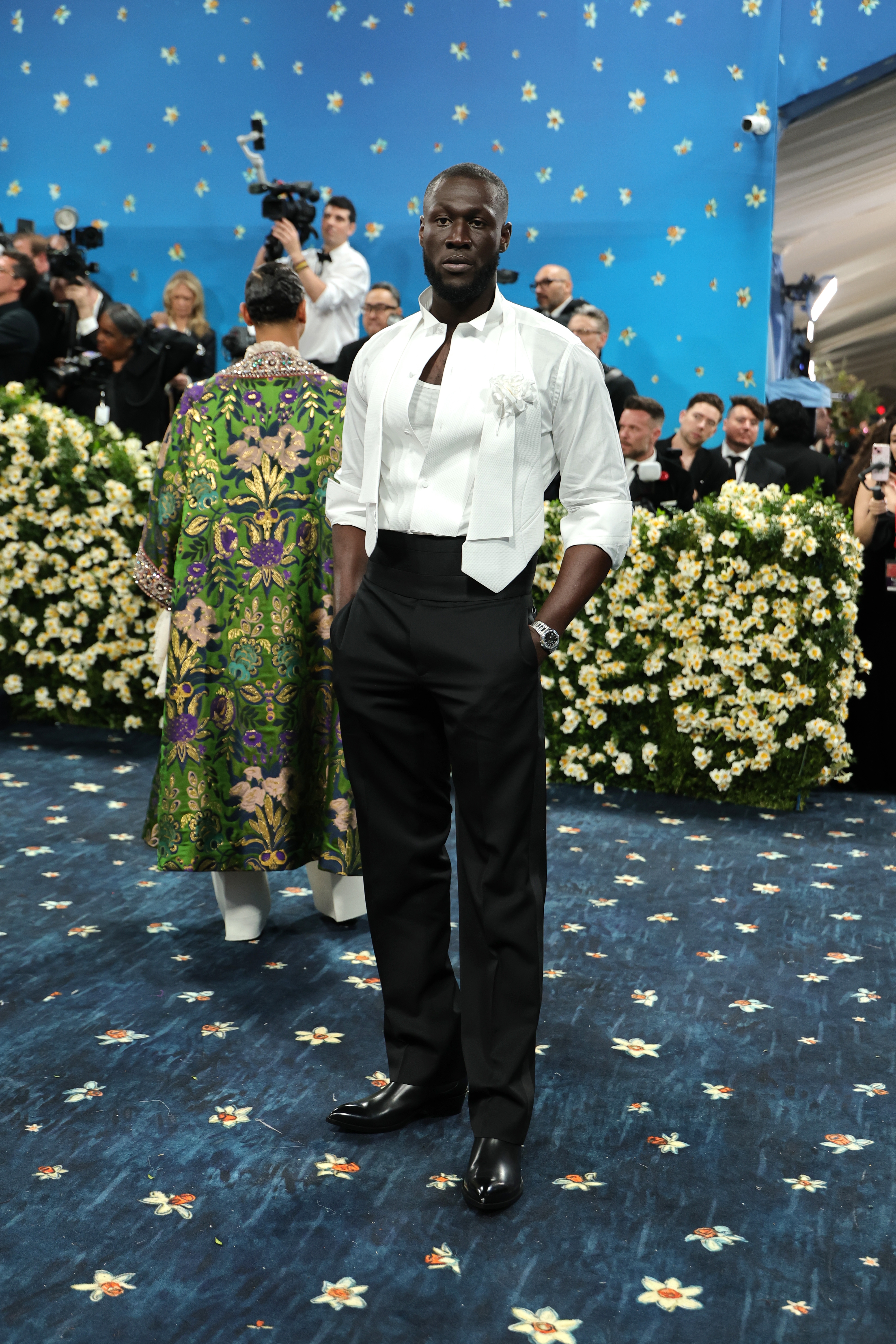 Person on a blue floral-patterned carpet, wearing a white shirt and vest with black pants. Cameras and attendees in the background