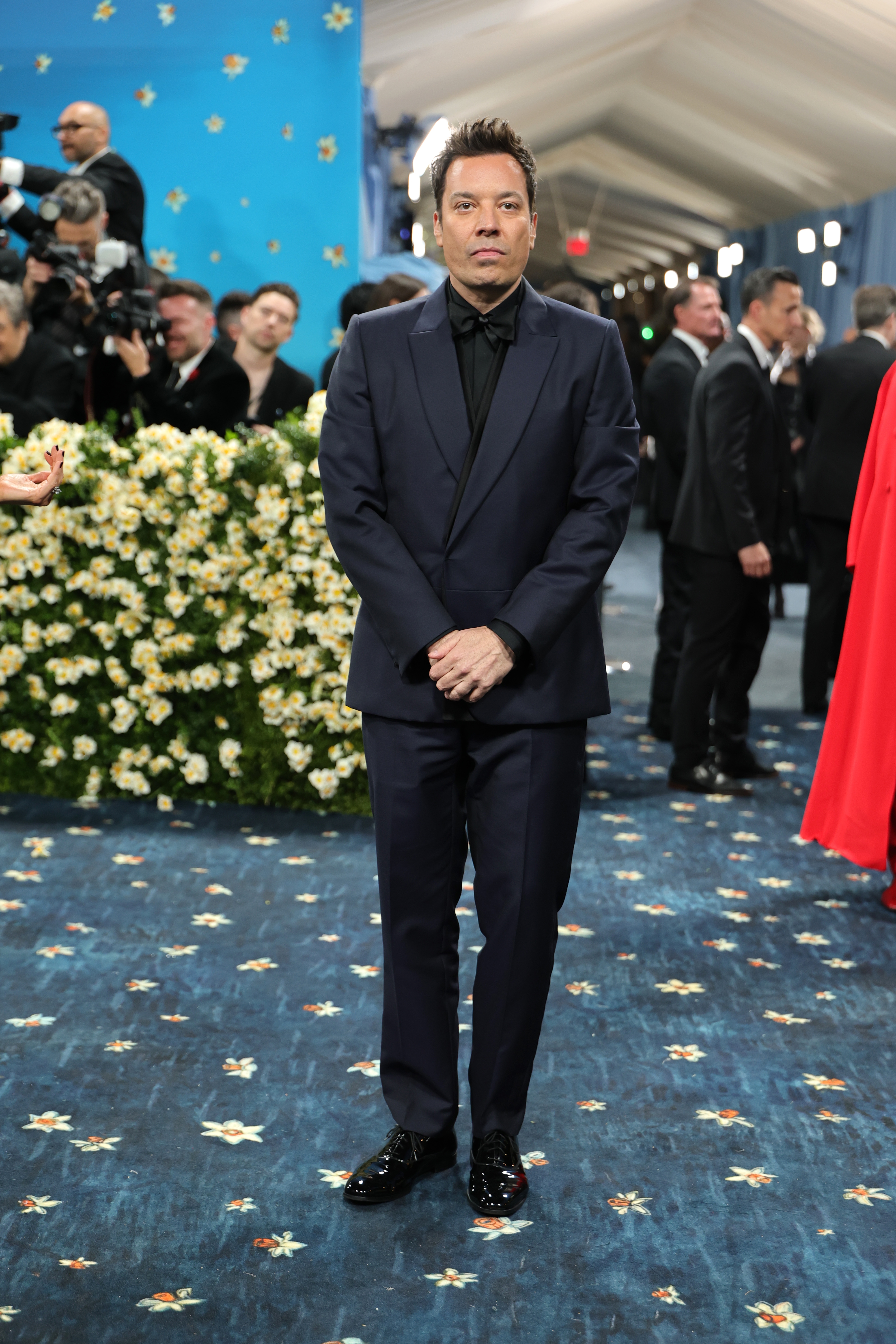 Man in formal dark suit standing on a blue-patterned carpet at a celebrity event, with photographers and guests in the background