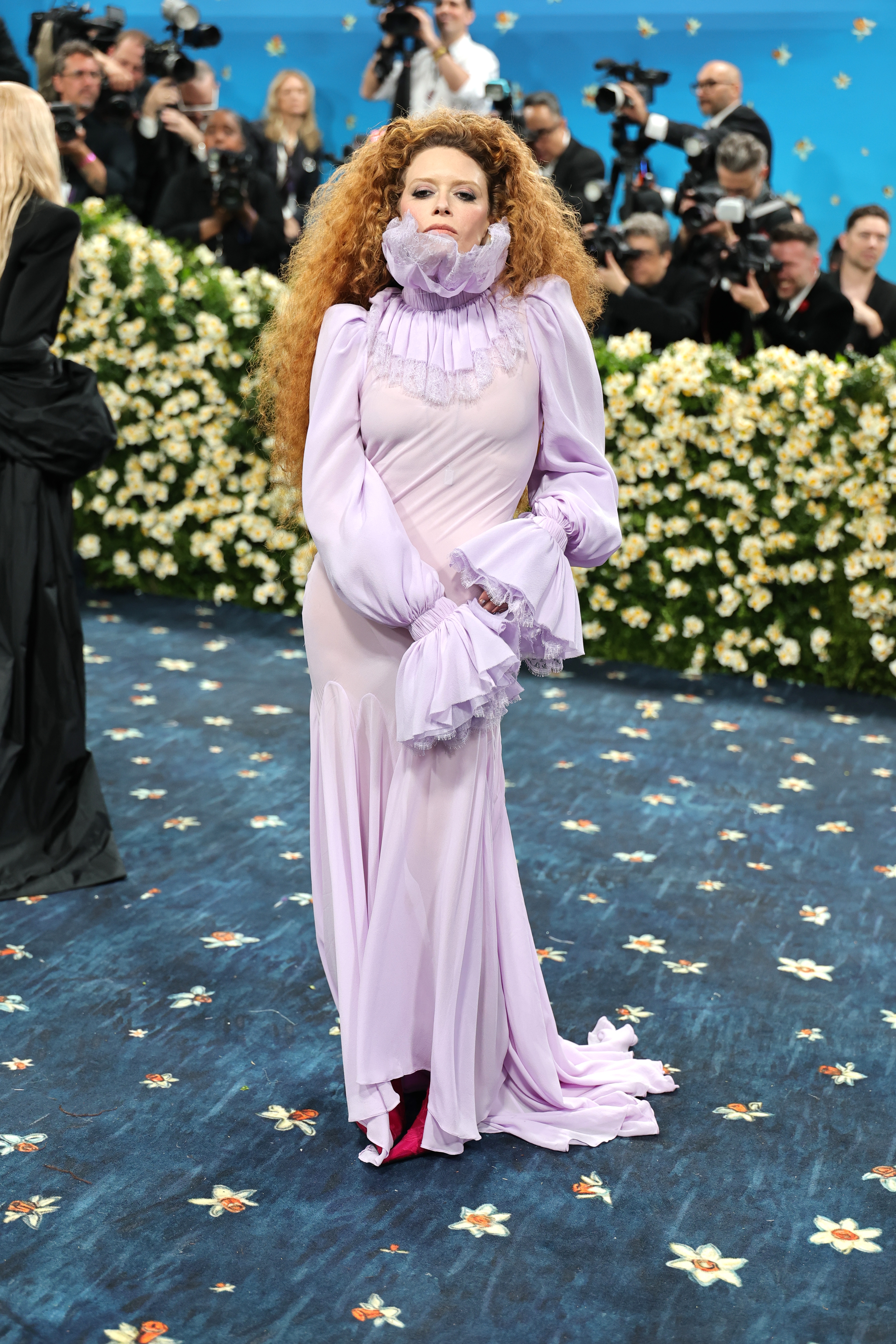 Person on a red carpet wearing a flowing gown with dramatic sleeves and a ruffled neckline, surrounded by photographers and floral decor