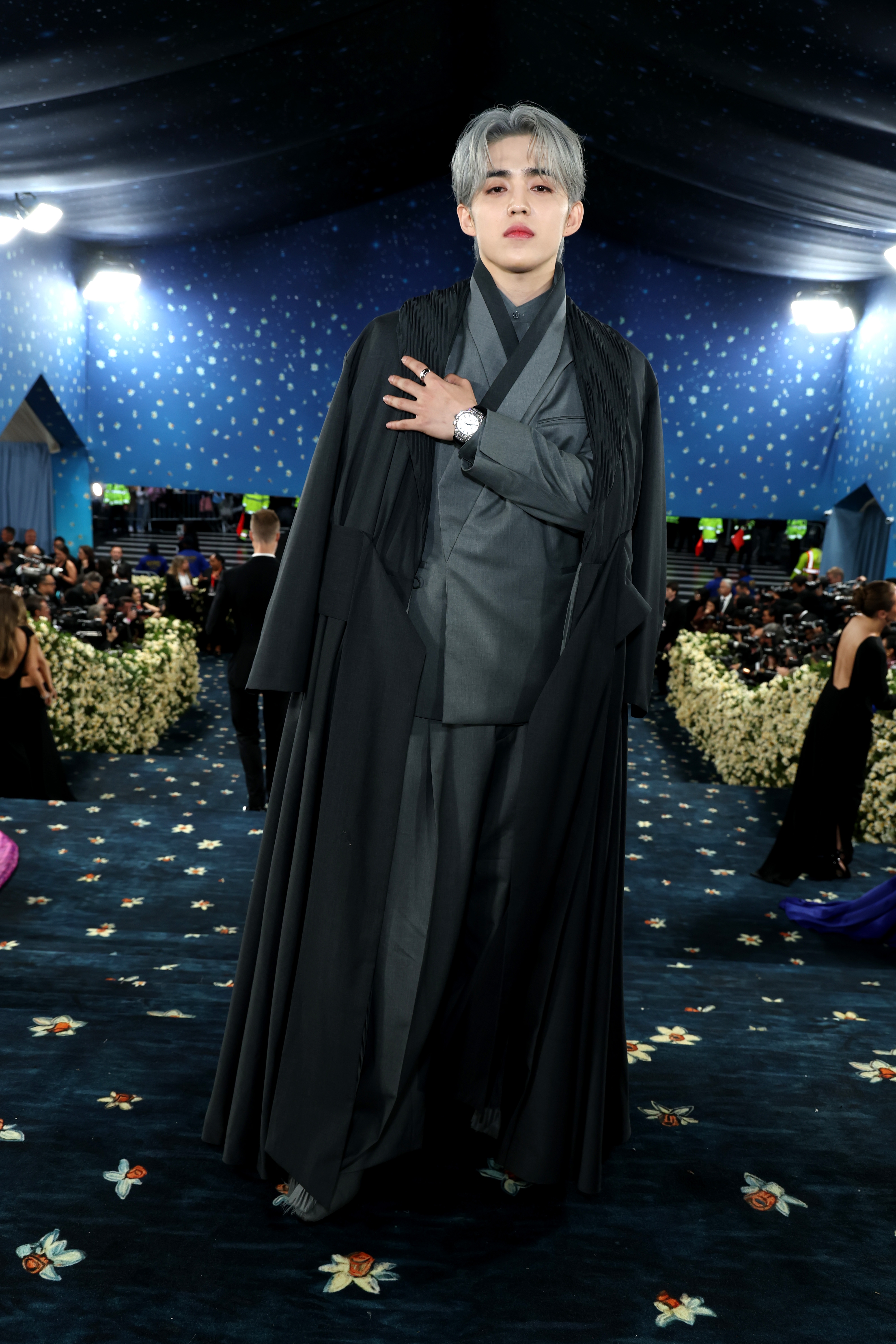 Person in an elegant black and gray layered outfit with long coat at a formal event, standing before a crowd and floral decorations