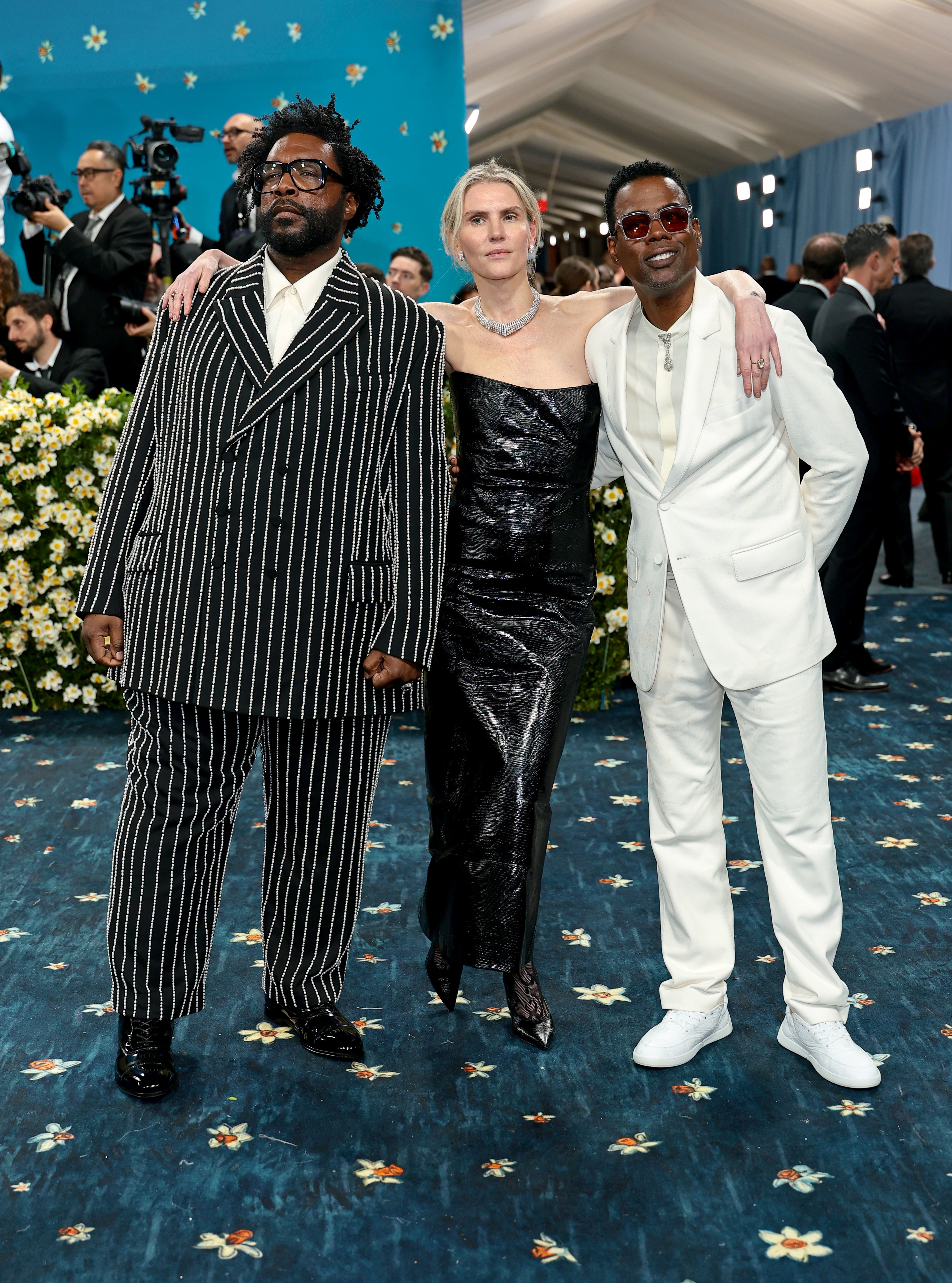 Three individuals pose together on a red carpet. The attire: pinstripe suit, metallic dress, and a white suit with sneakers