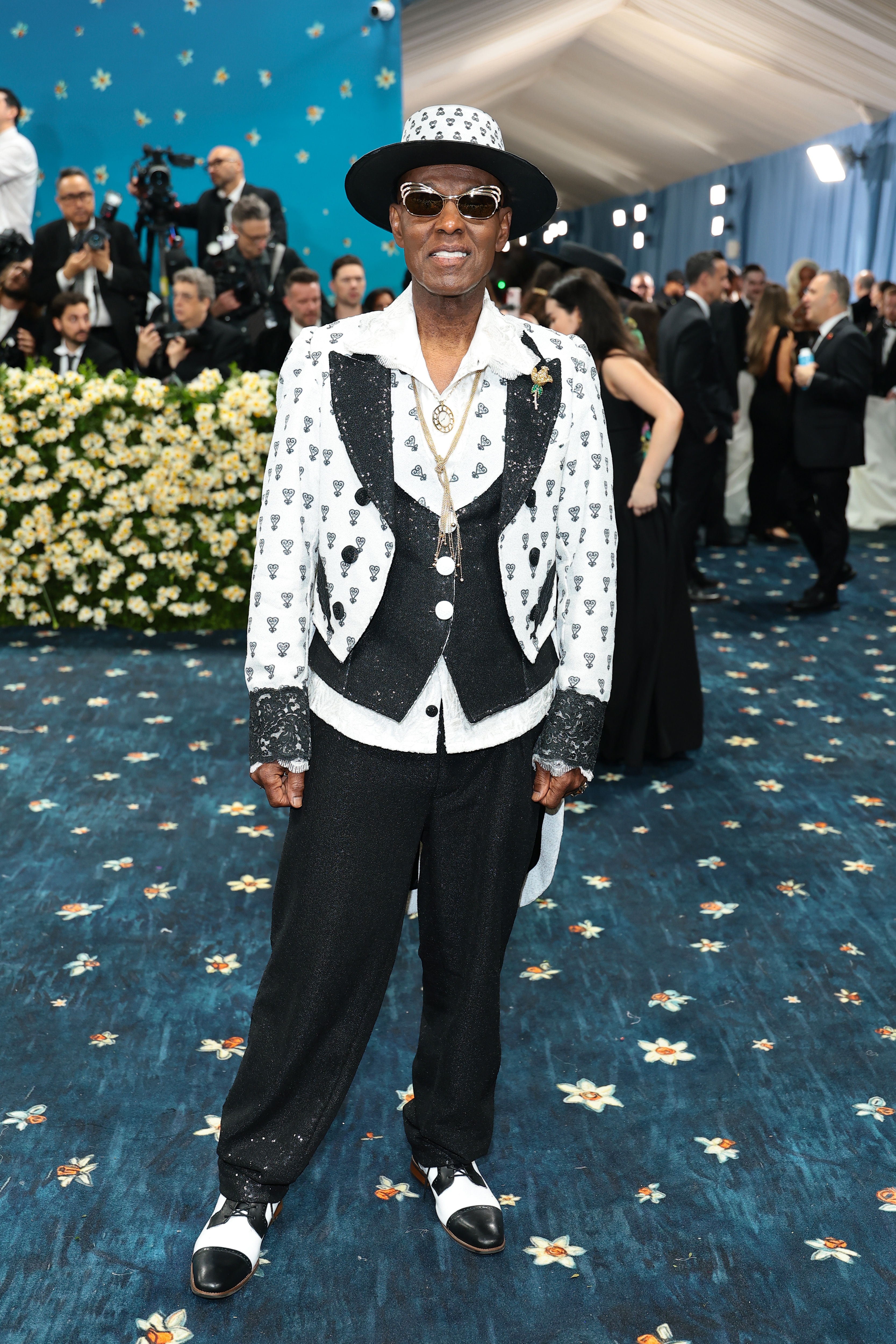 Person in a tuxedo with a heart pattern, wearing a fedora and sunglasses, on a formal event carpet with a floral backdrop and crowd