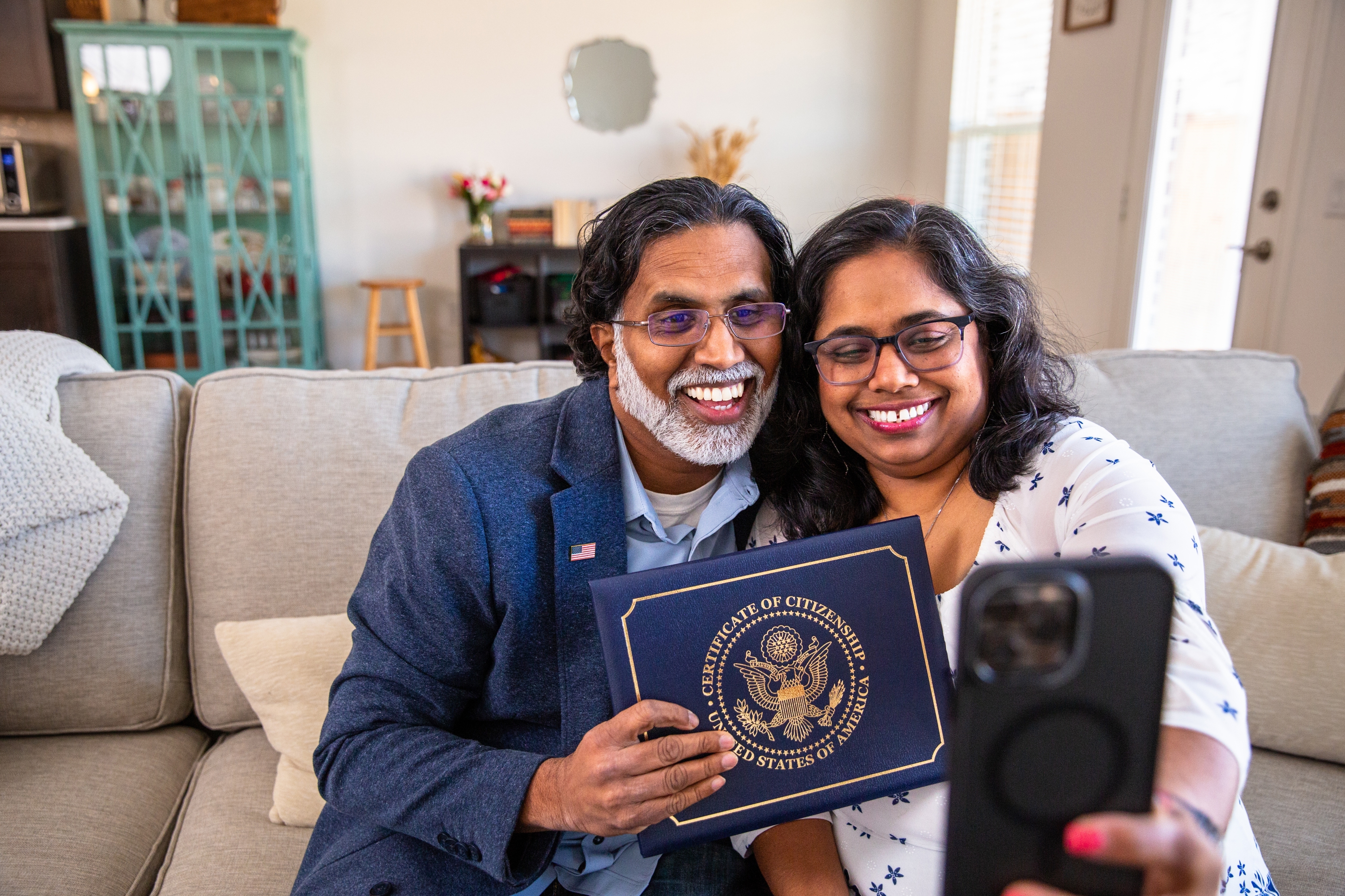 Two people sitting on a couch, smiling and taking a selfie. One person holds a Certificate of Citizenship