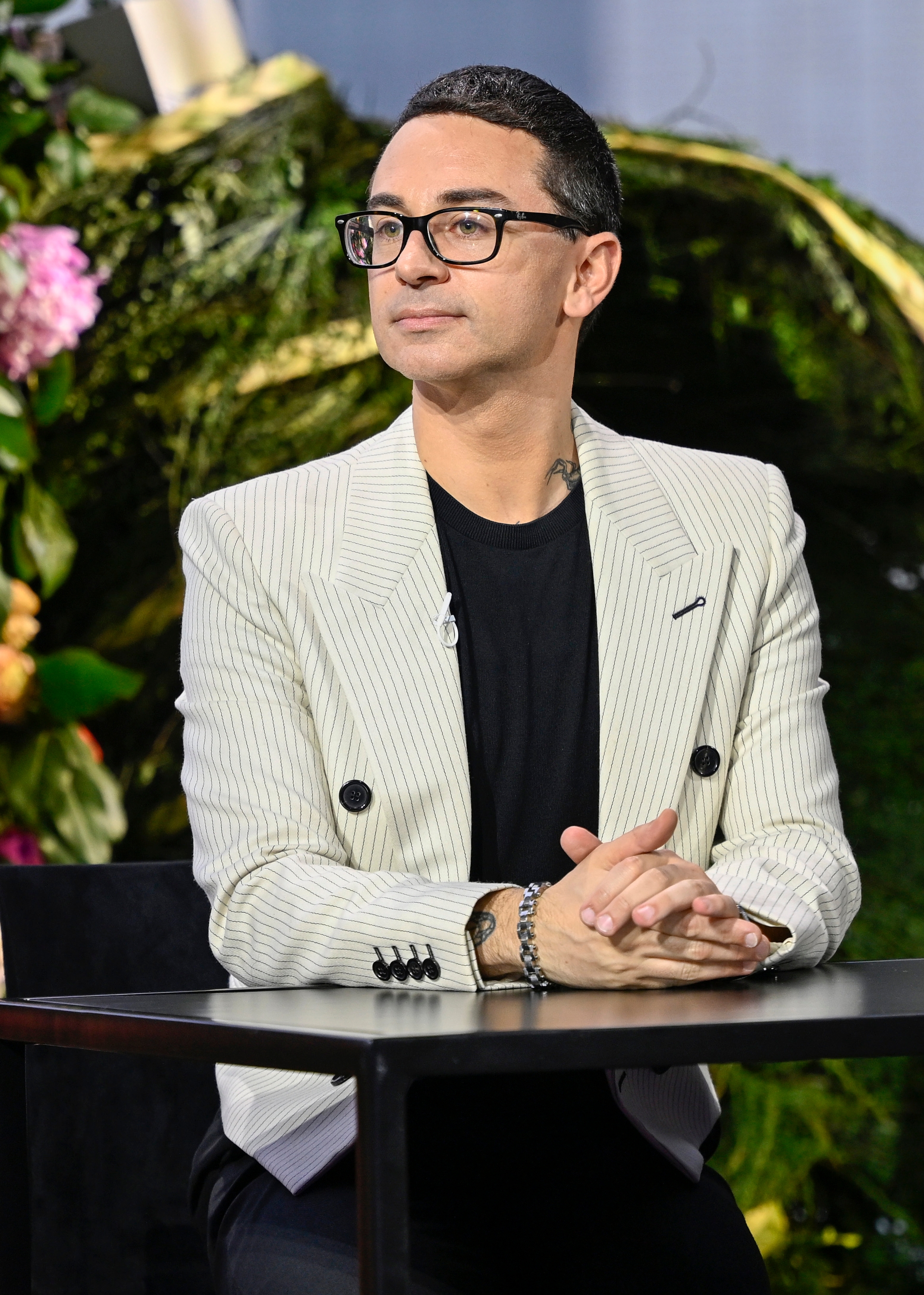 Christian Siriano wearing a pinstriped blazer and glasses is seated at a table, hands clasped, in an event setting with a floral-themed backdrop
