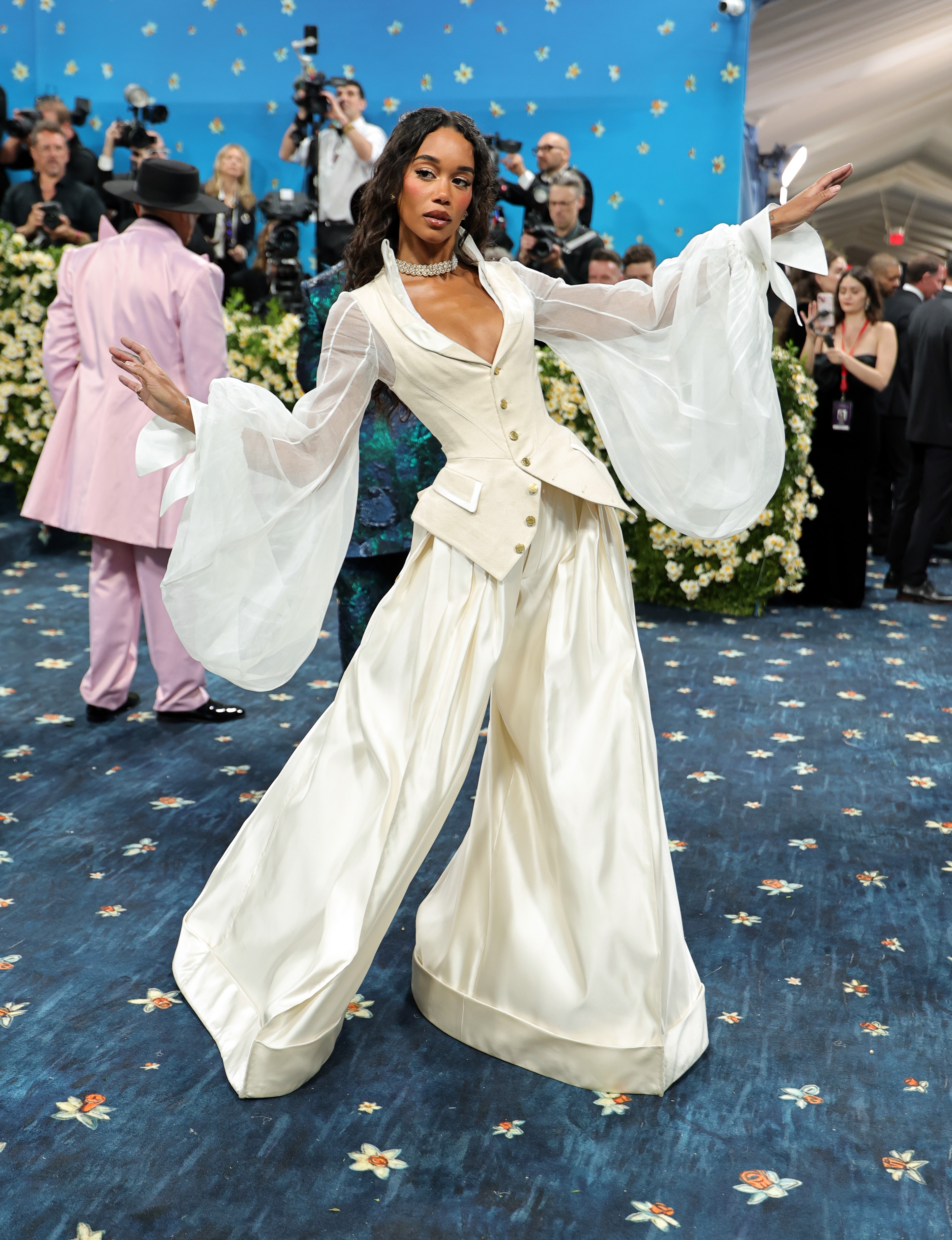 Person poses on a floral-themed carpet in an elegant, flowing outfit with sheer sleeves, surrounded by photographers and onlookers