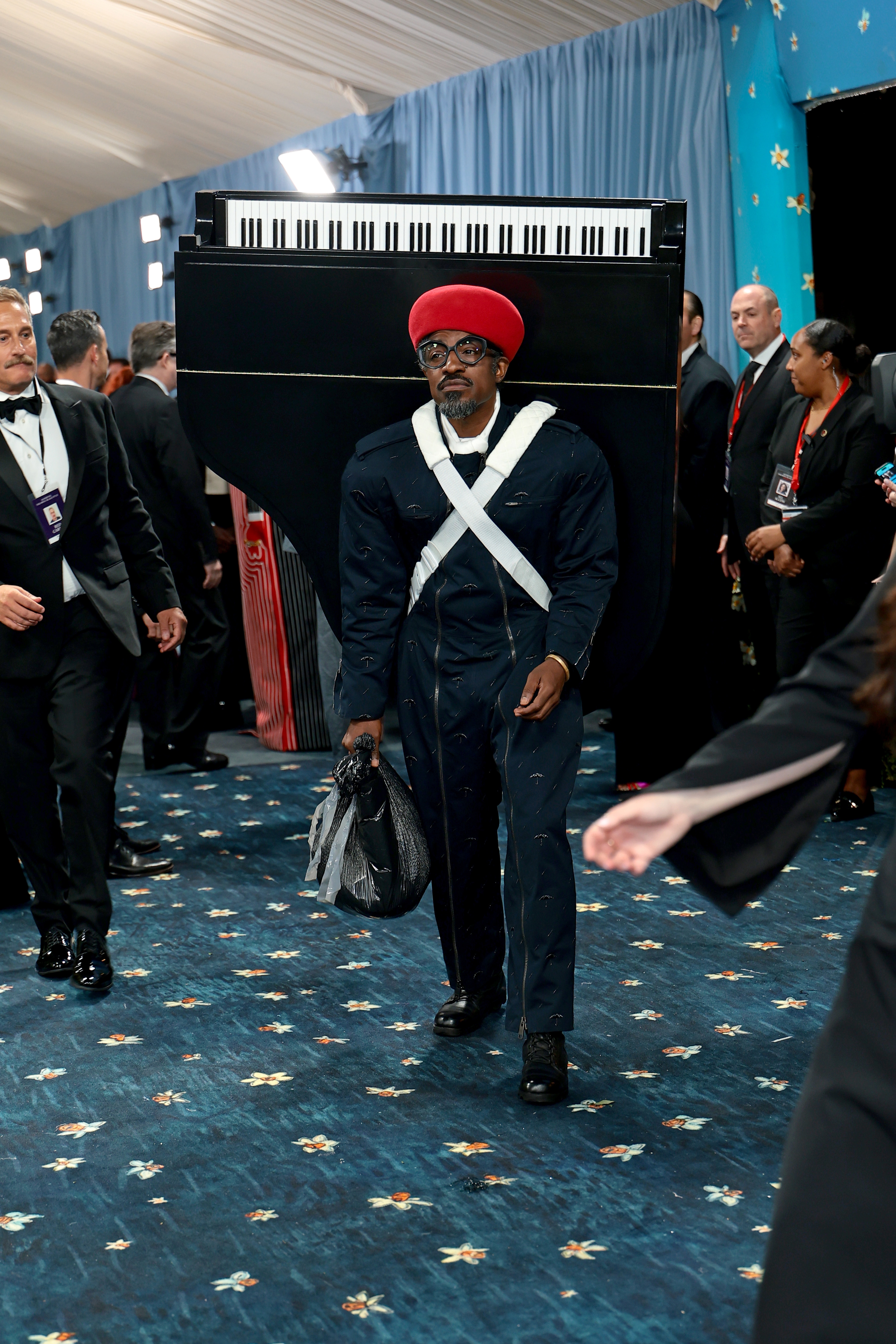 Person in a unique outfit resembling a piano with a keyboard design and a red hat, carrying a black bag, walking at an event