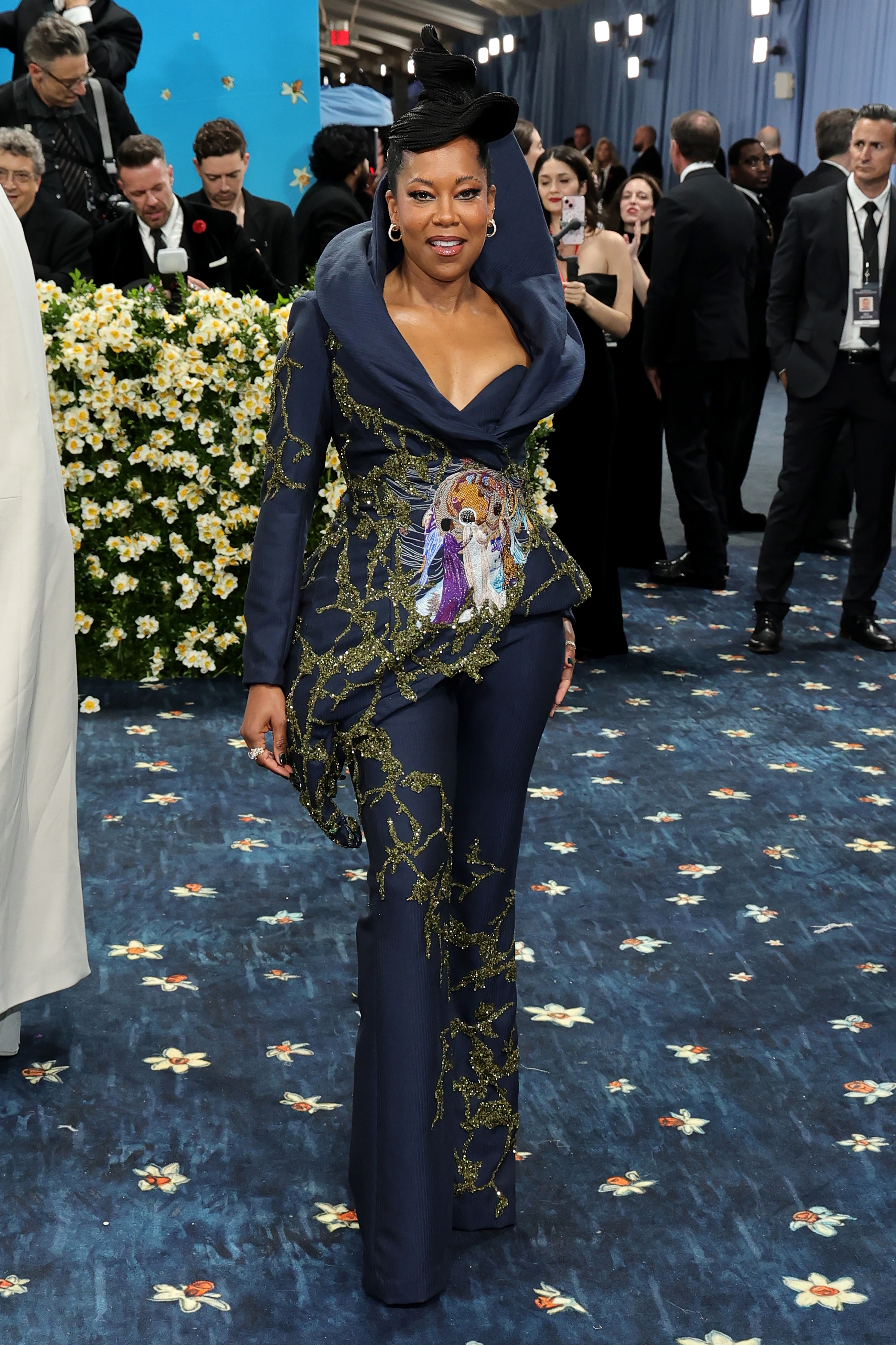 Person in a stylish embroidered suit poses on a red carpet, surrounded by event attendees