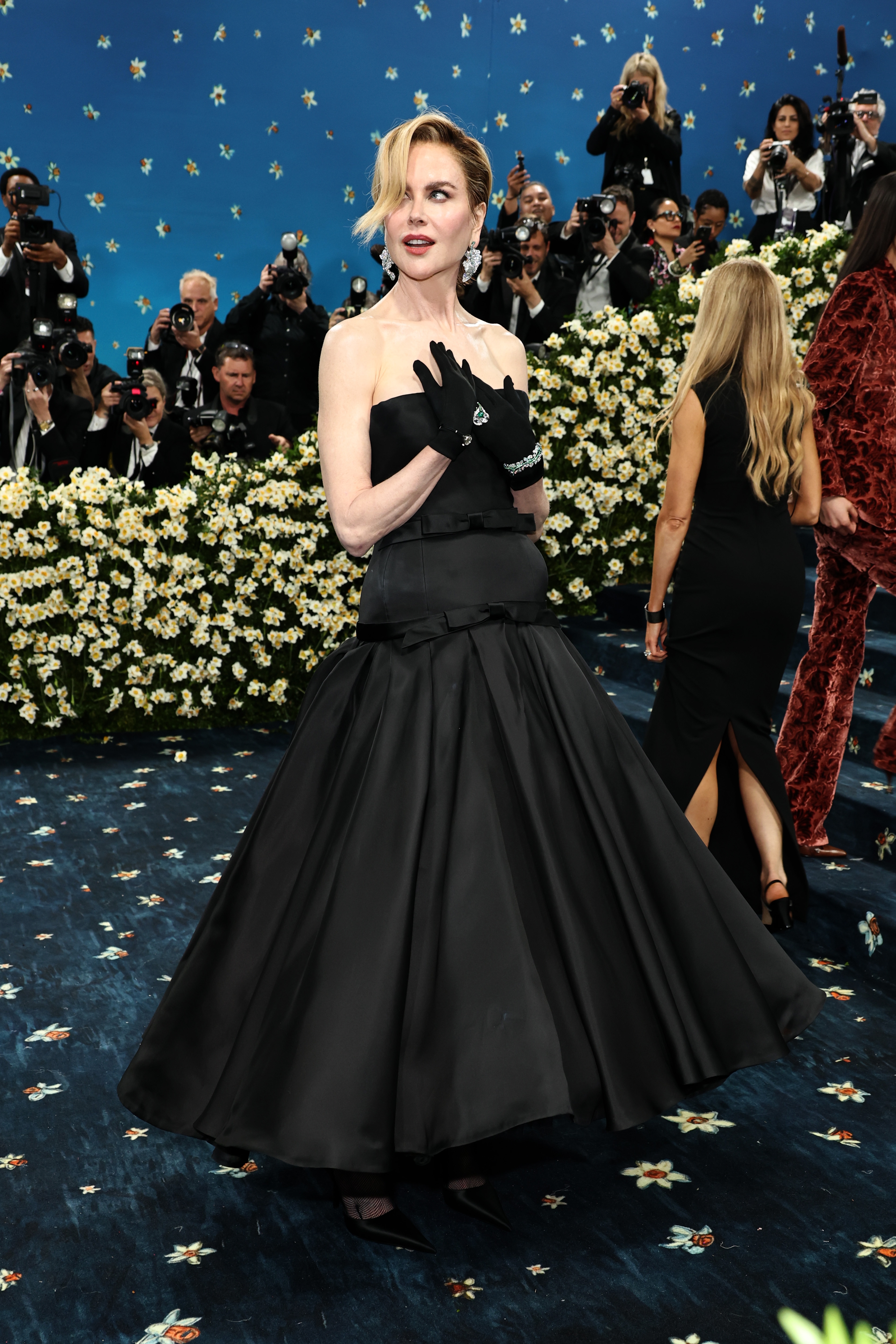 Actor in elegant black strapless gown with full skirt, black gloves posing on a floral-themed red carpet event. Photographers and attendees in background