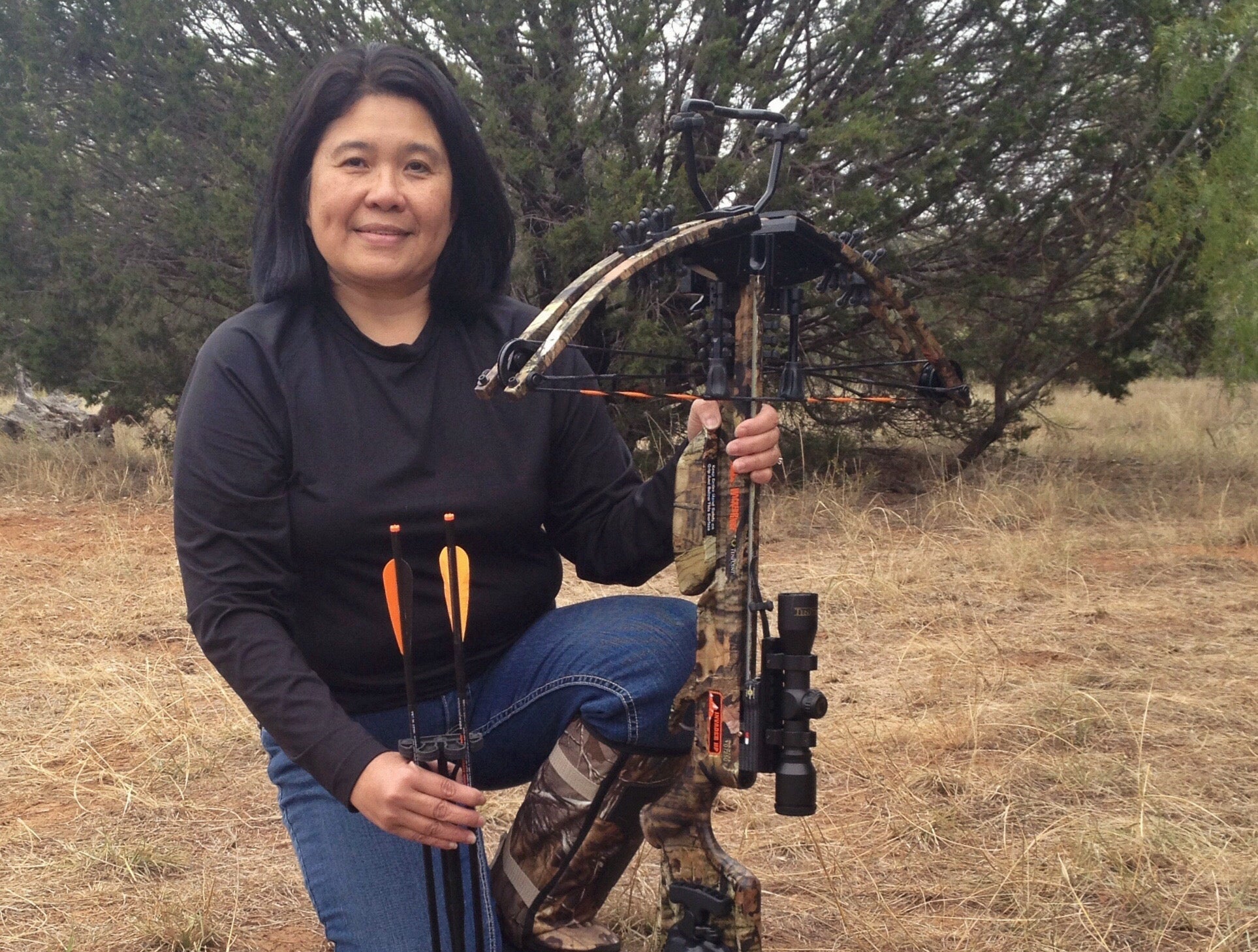 Person kneels in field with bow, holding arrows next to a wild turkey. Trees visible in the background